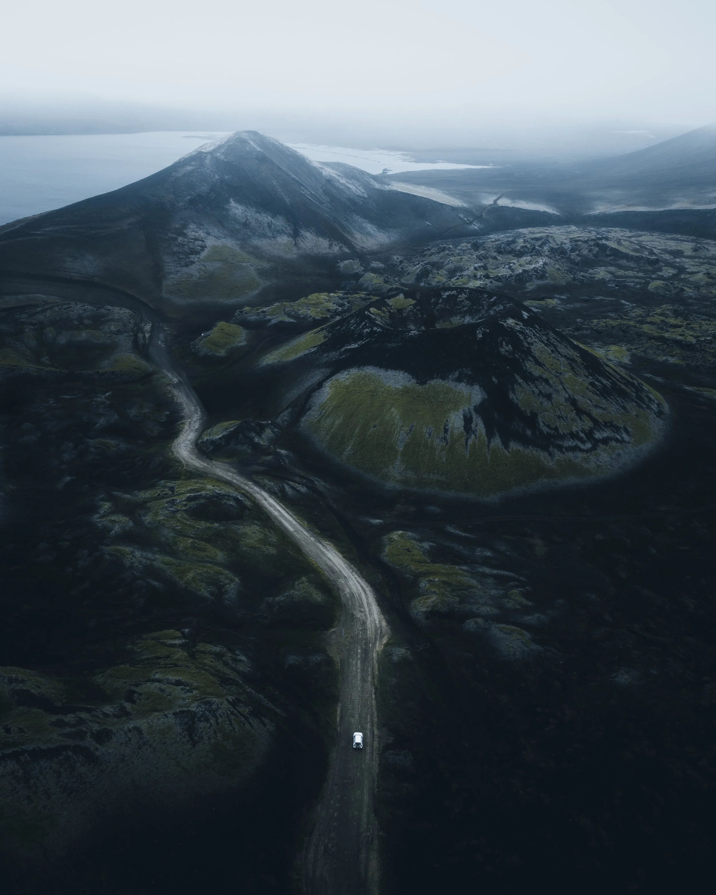 Aerial view of a winding dirt road with a white vehicle driving on it, surrounded by dark green and moss-covered rocky hills, with a mountain in the background and a body of water in the distant horizon.