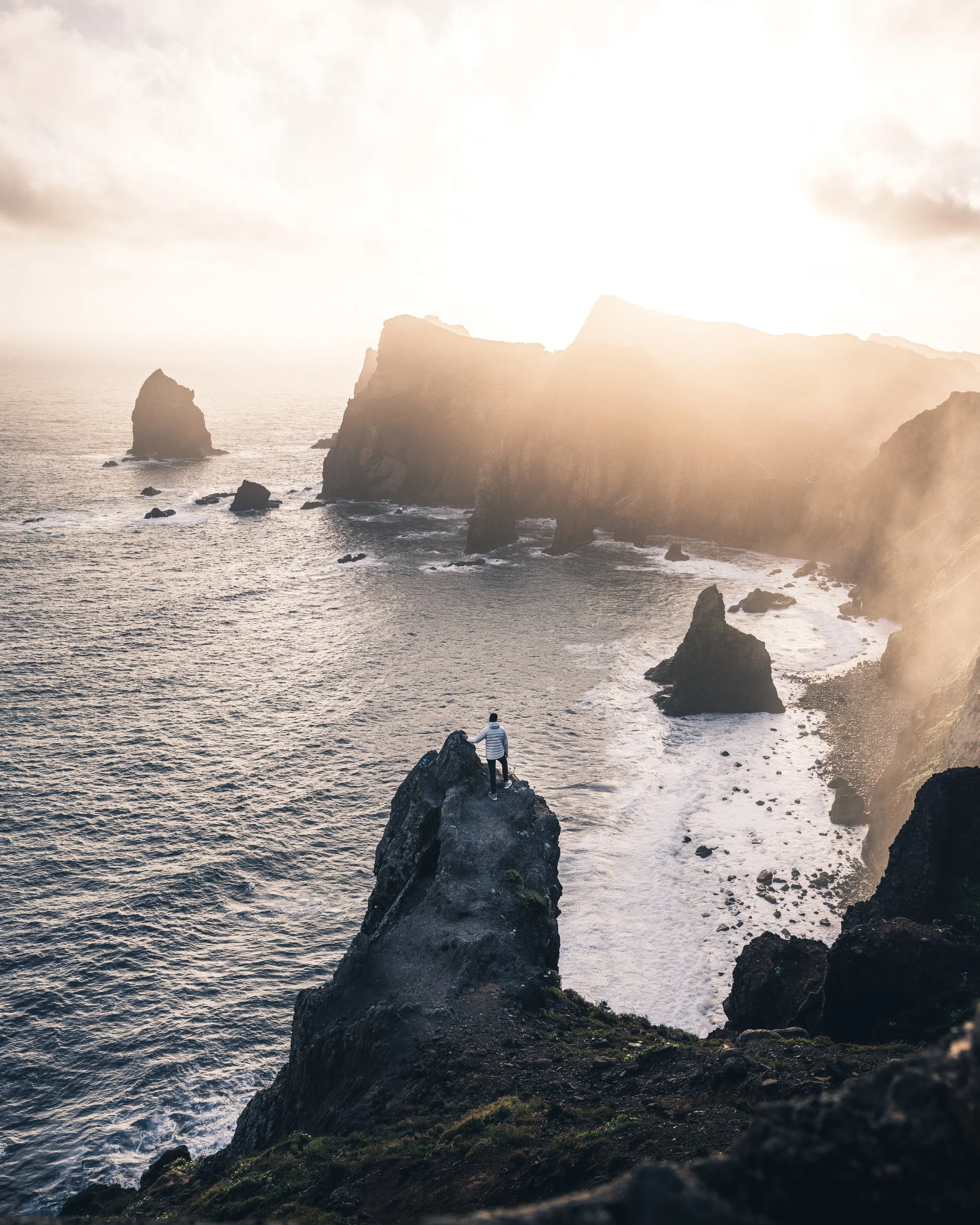 Person standing on a rock at the edge of the ocean, with large cliffs and rock formations in the background, during sunset or sunrise.