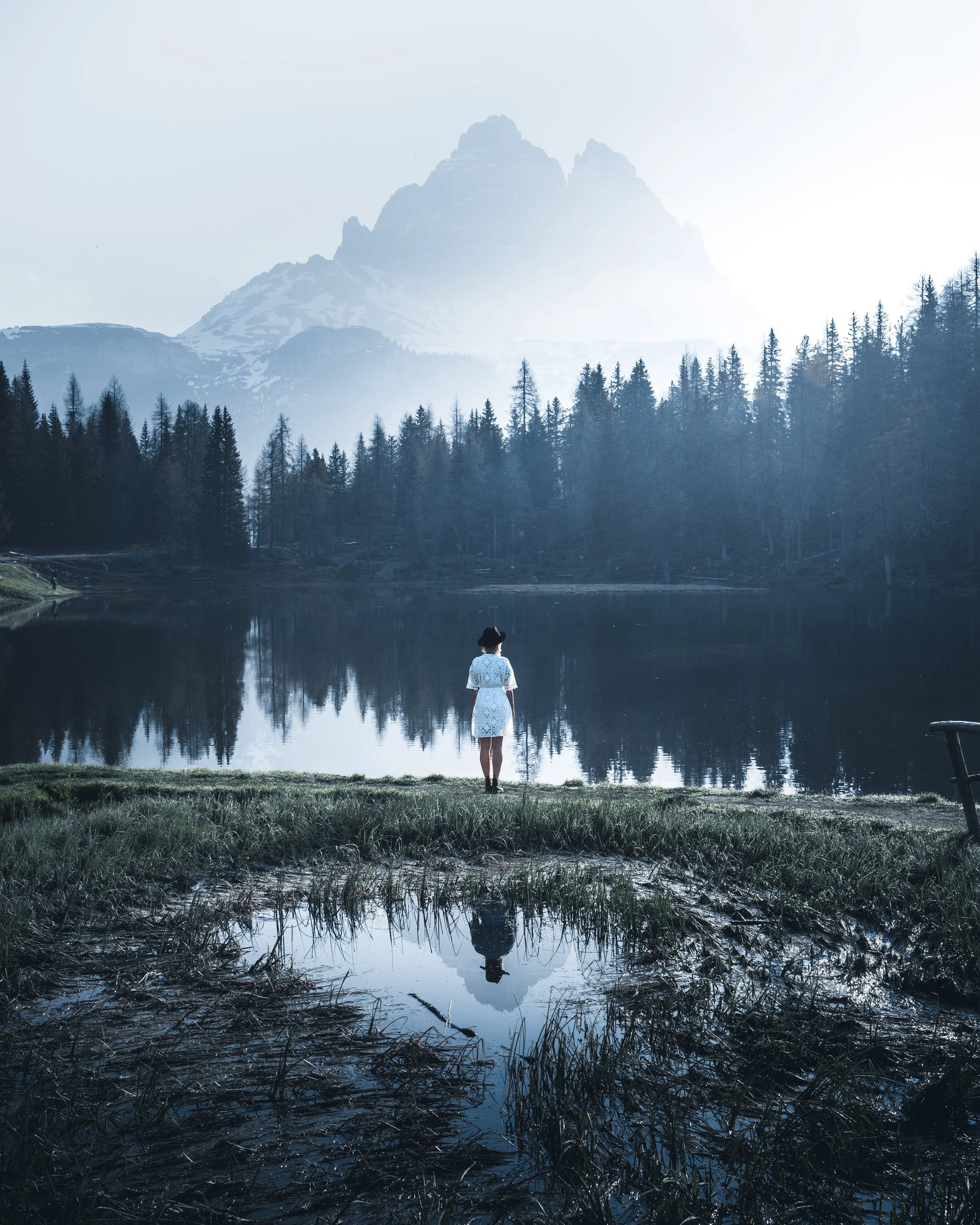 A woman in a white dress and black hat stands at the edge of a lake, with a mountain range and trees in the background, and her reflection visible in the water.