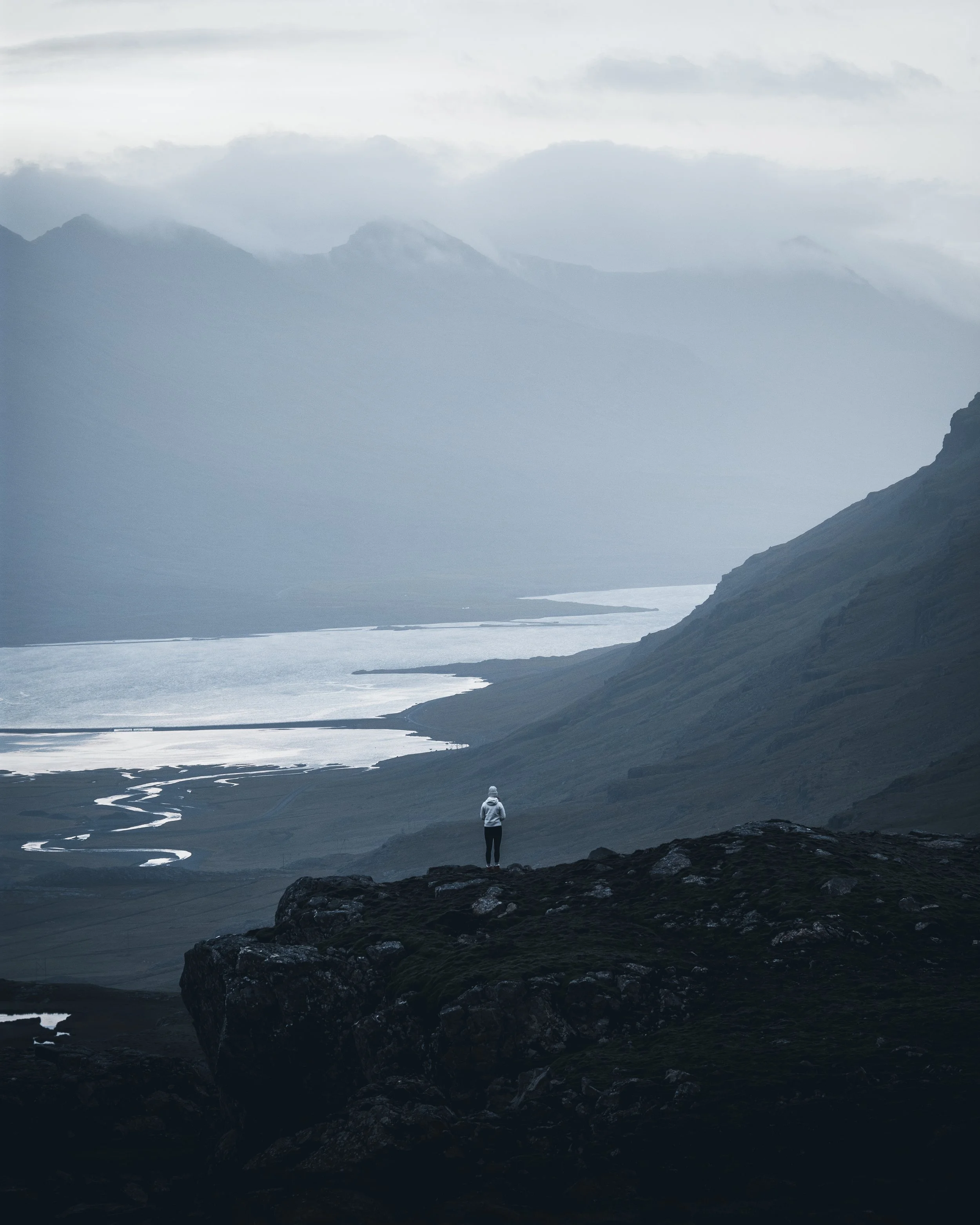 Person standing on rocky terrain overlooking a vast, misty landscape with winding water paths, mountains in the background, and cloudy sky.