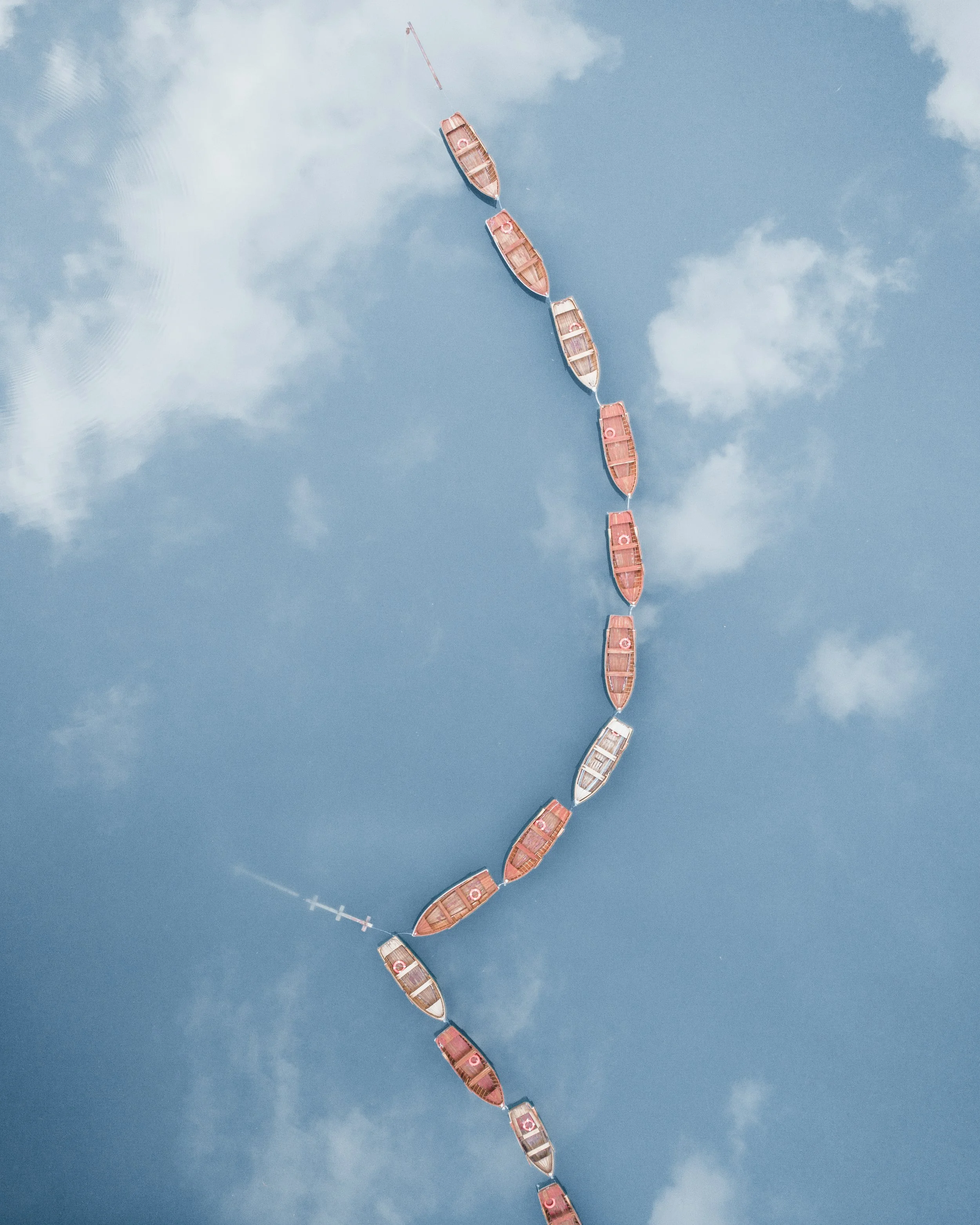 Aerial view of several small boats tethered in a line on calm water with a blue sky and scattered clouds.