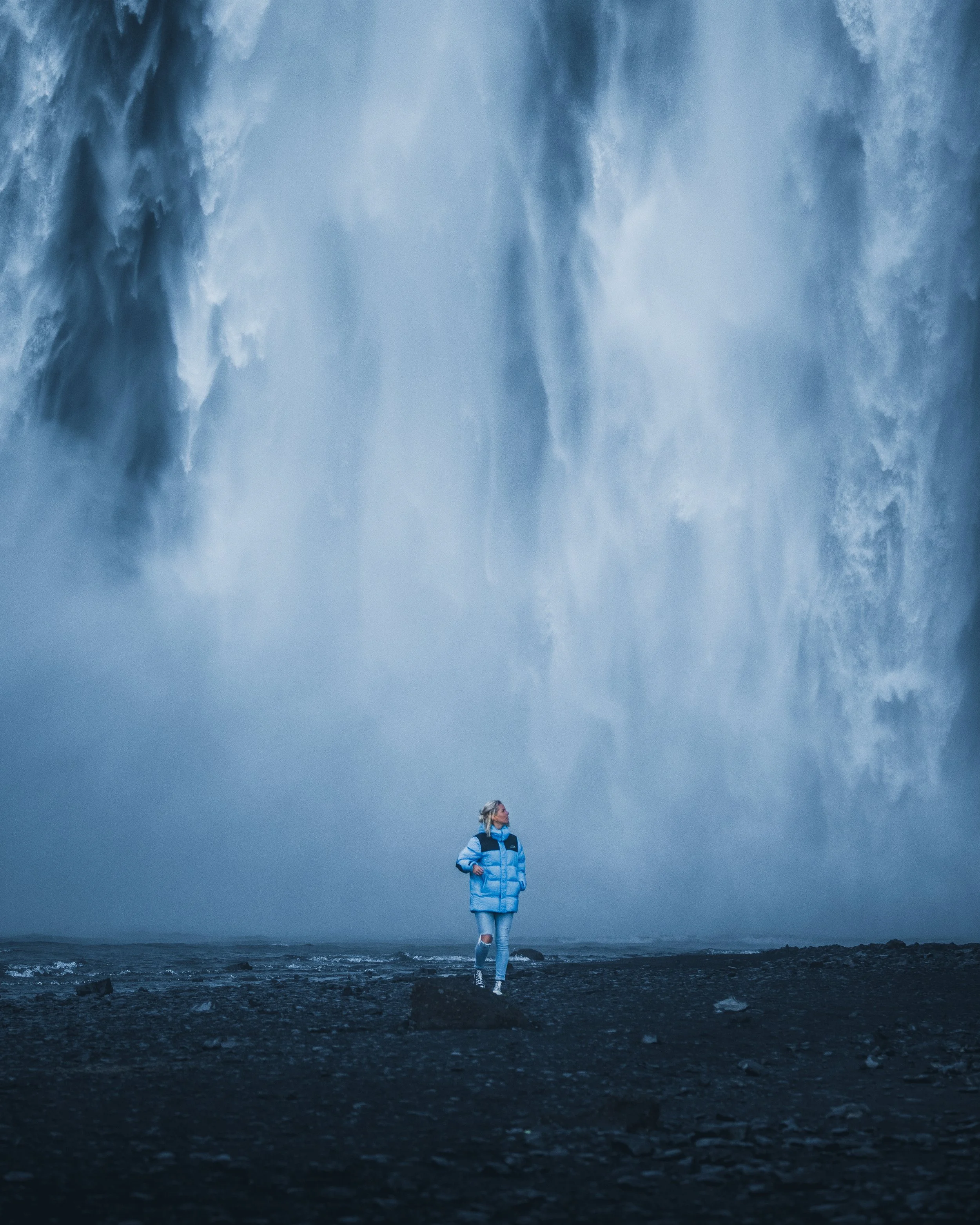 A woman standing on rocky terrain near the ocean with a massive waterfall cascading down behind her.