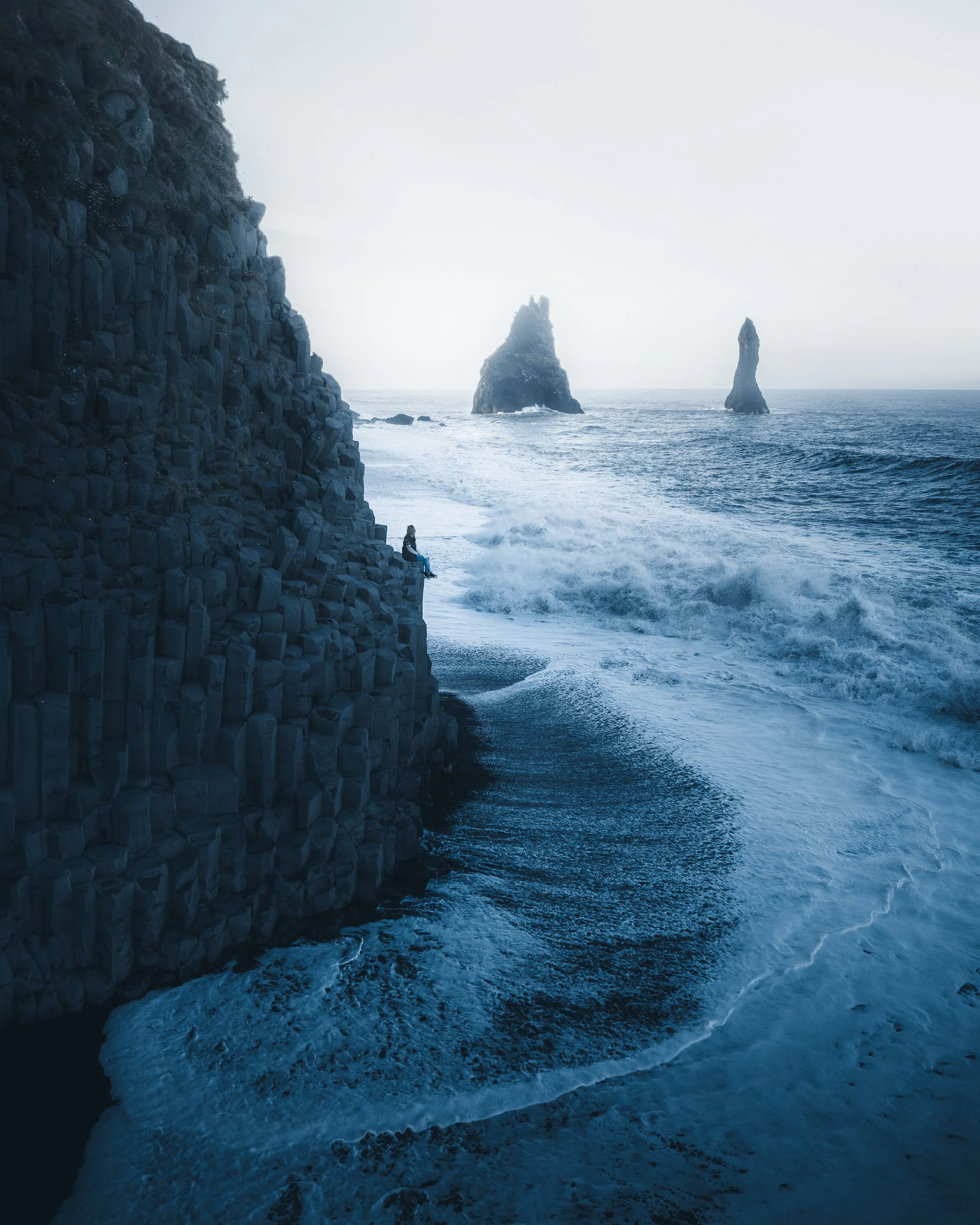 A person sitting on rocks near the shoreline, facing the ocean, with two large sea stacks in the background during foggy weather.