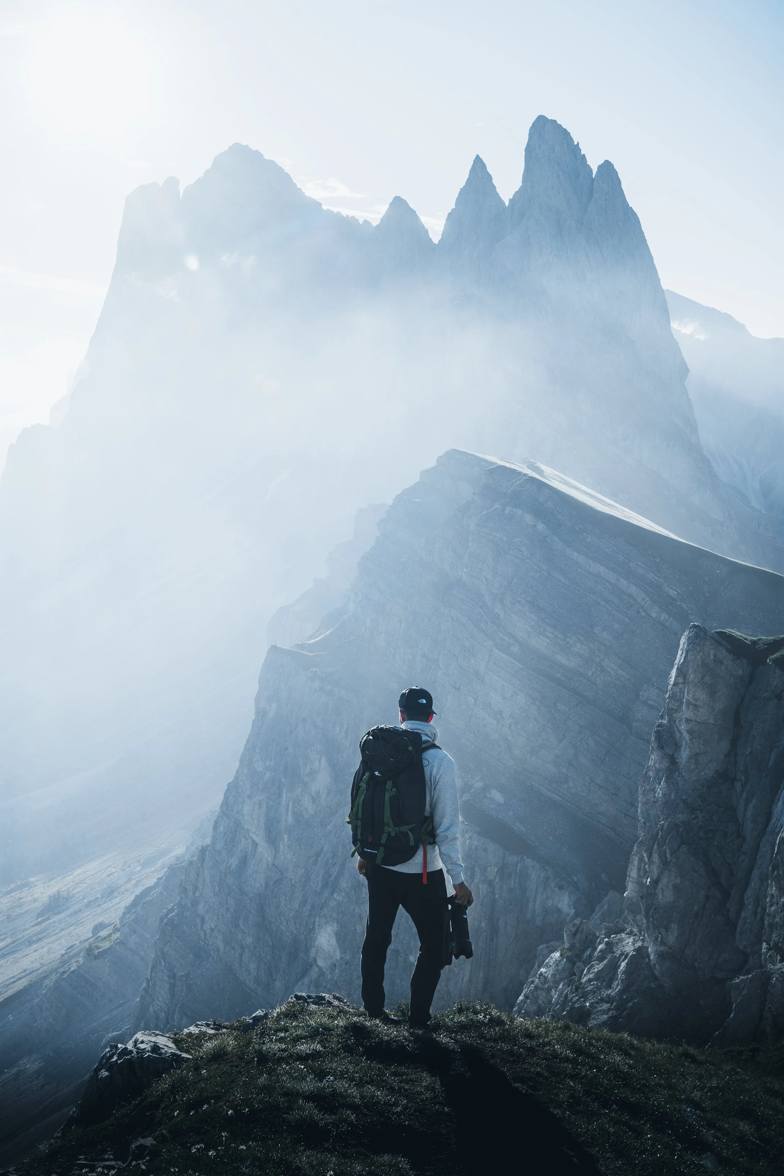 A person with a backpack and hat standing on a rocky outcrop, looking at a mountain range shrouded in mist and clouds.