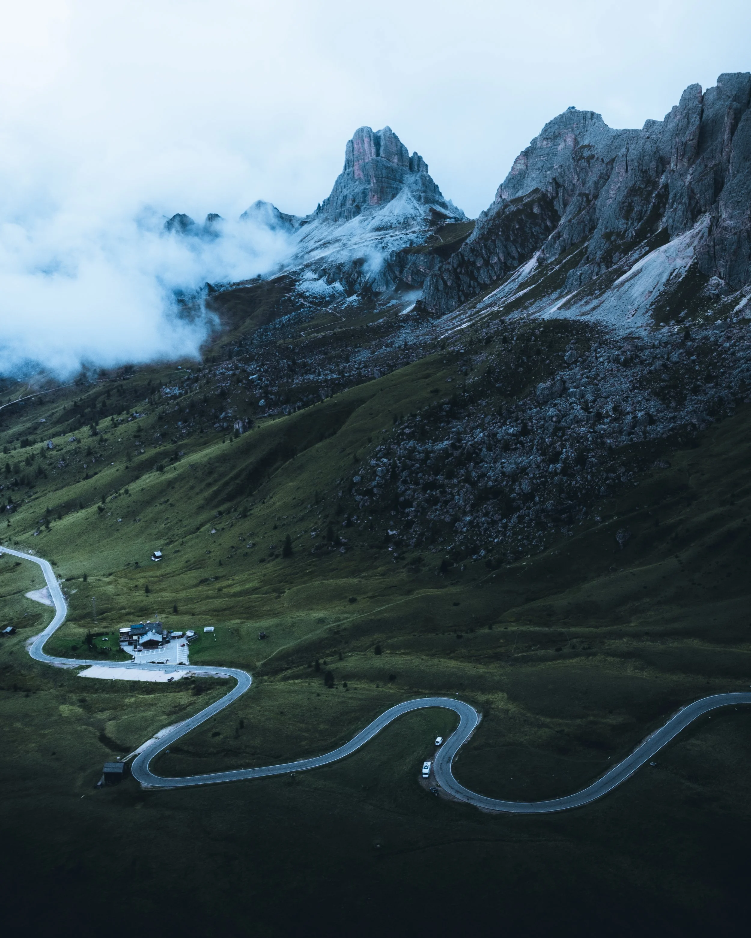 Mountain landscape with a winding road and a green hillside, fog around the peaks.