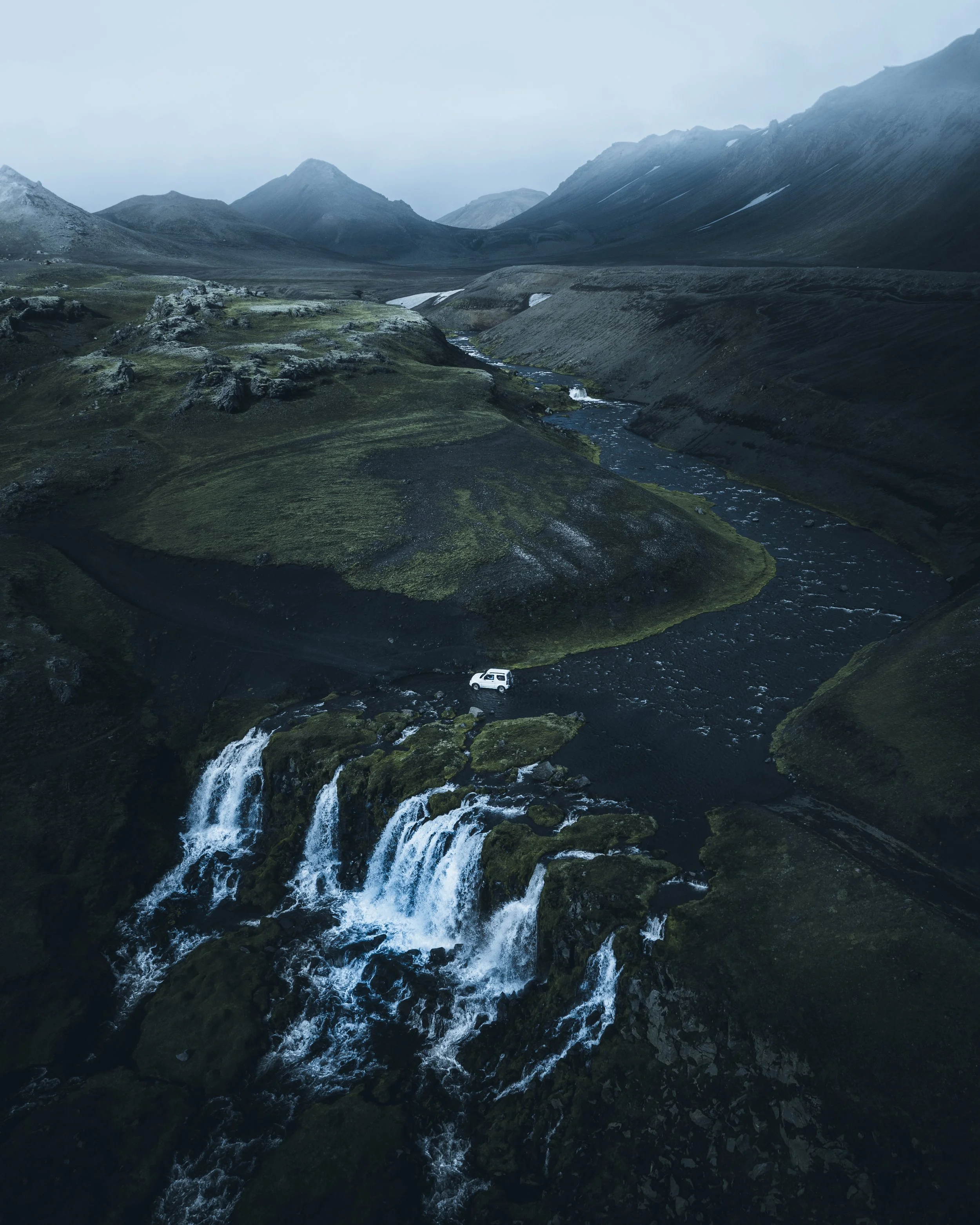 A rugged mountain landscape with a winding river and a small white van crossing a stream.