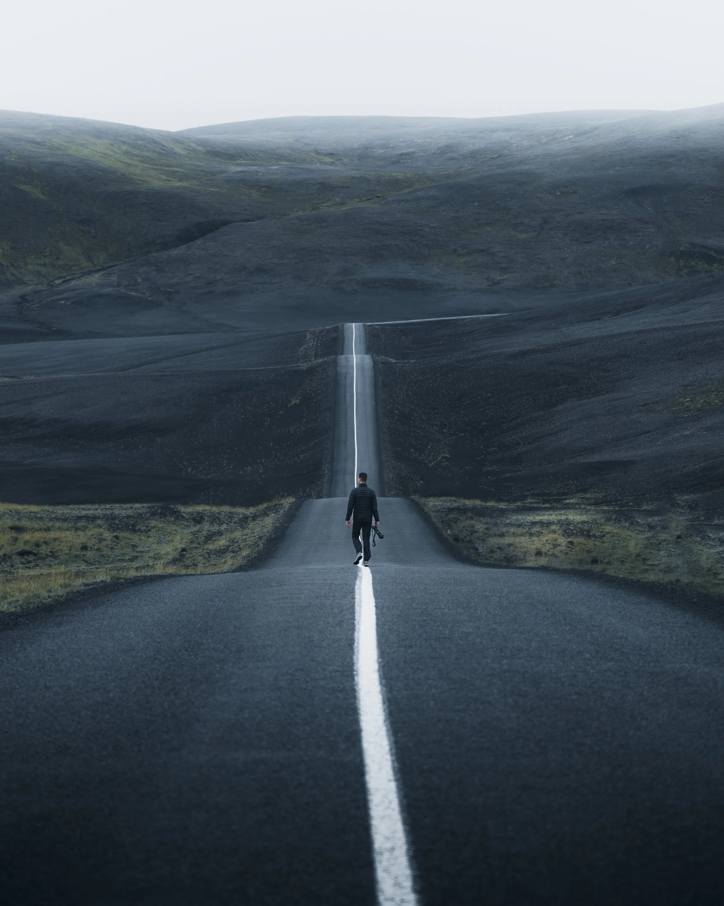A person walking on a narrow road in a desolate landscape with dark rolling hills and an overcast sky, holding a camera in one hand.