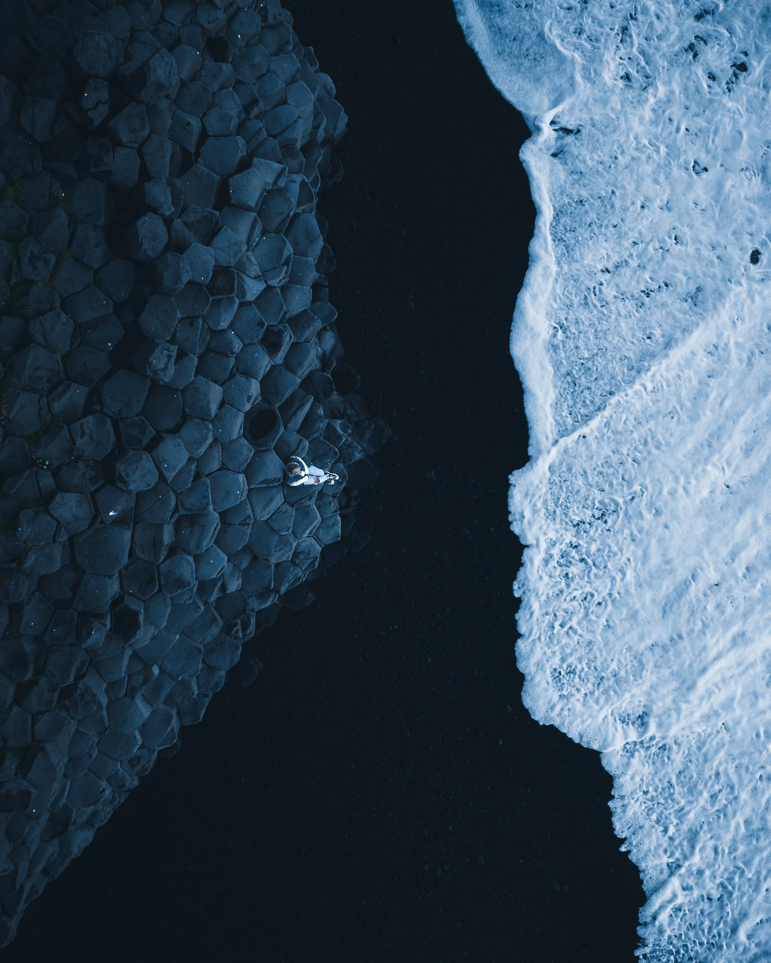 A drone view of a shoreline showing dark rocks on the left and white foamy waves on the right.