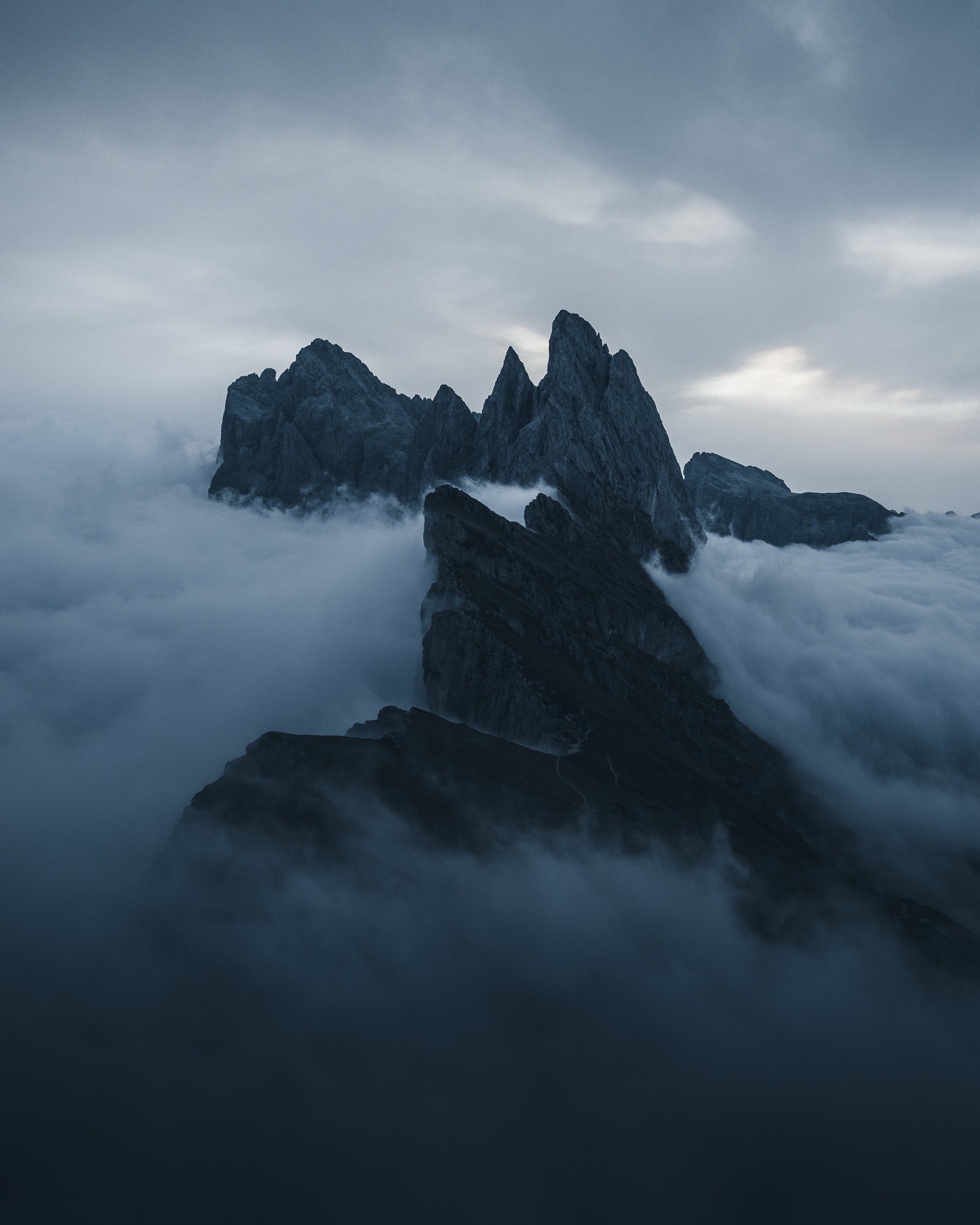 A rugged mountain range with sharp peaks, shrouded in mist and low-hanging clouds, under a cloudy sky.