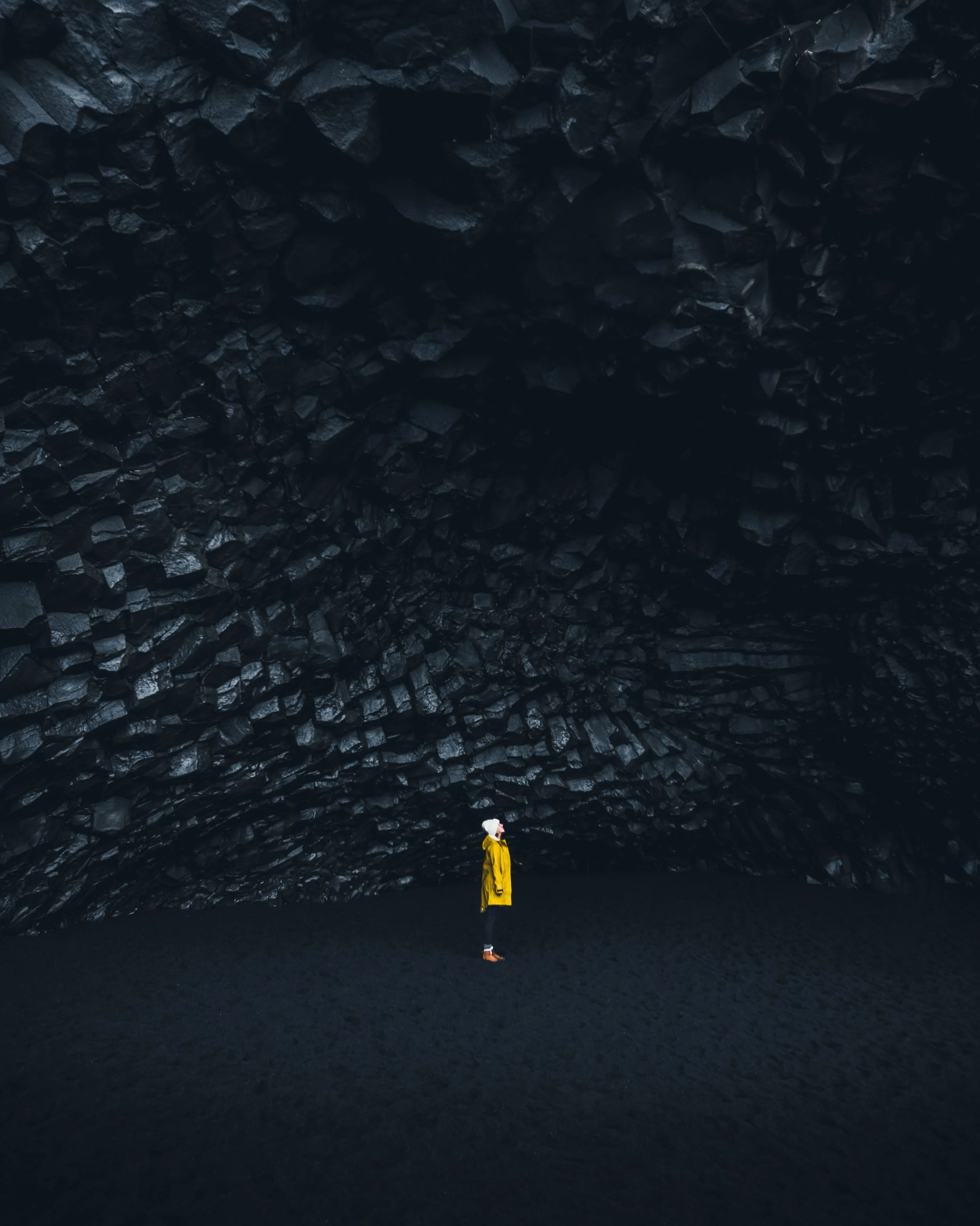 A person in a yellow jacket and white hat stands inside a dark volcanic cave with black rocks lining the ceiling and floor.