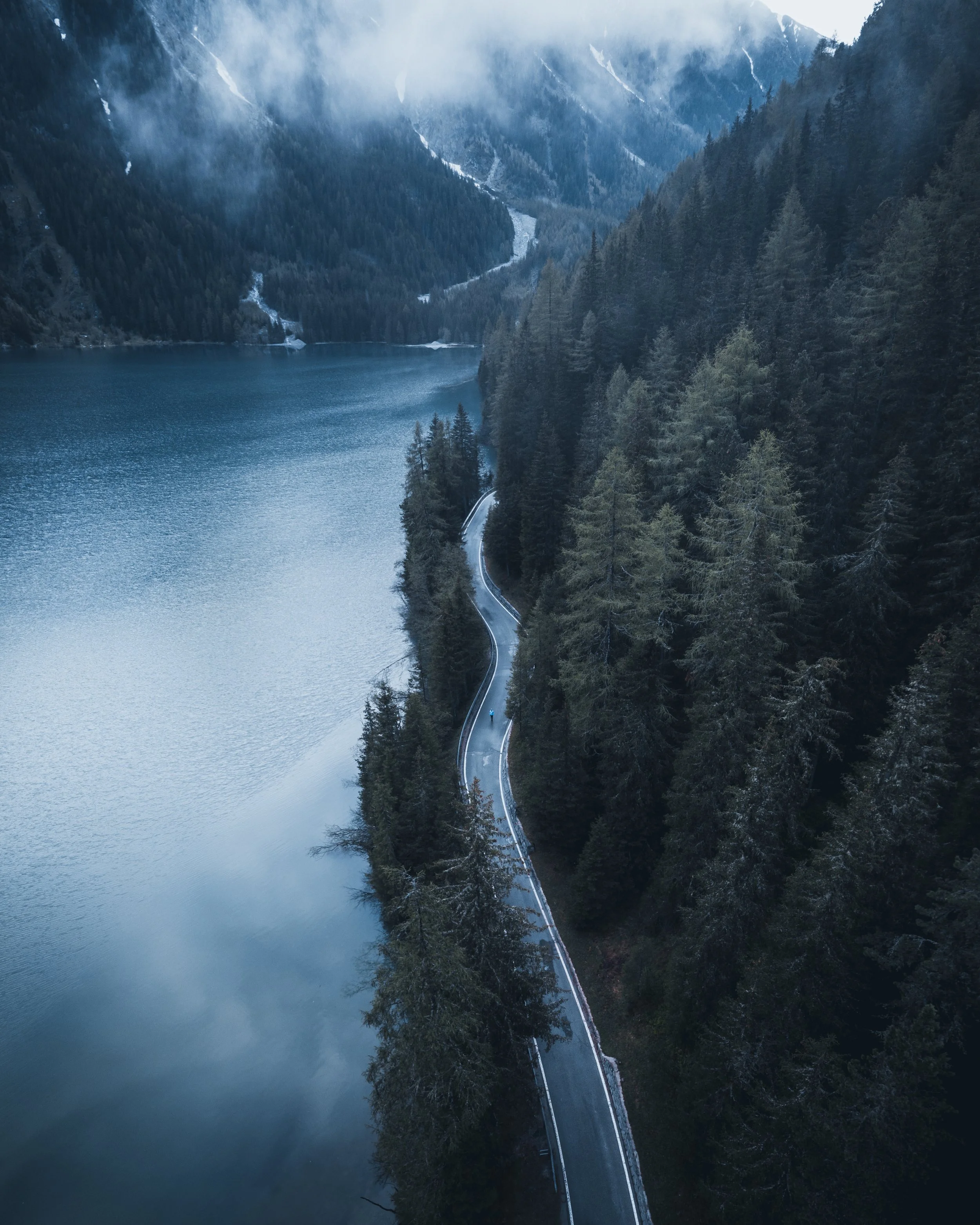 A winding road runs along a forested hillside beside a large mountain lake, with snow-capped mountains and mist in the background.