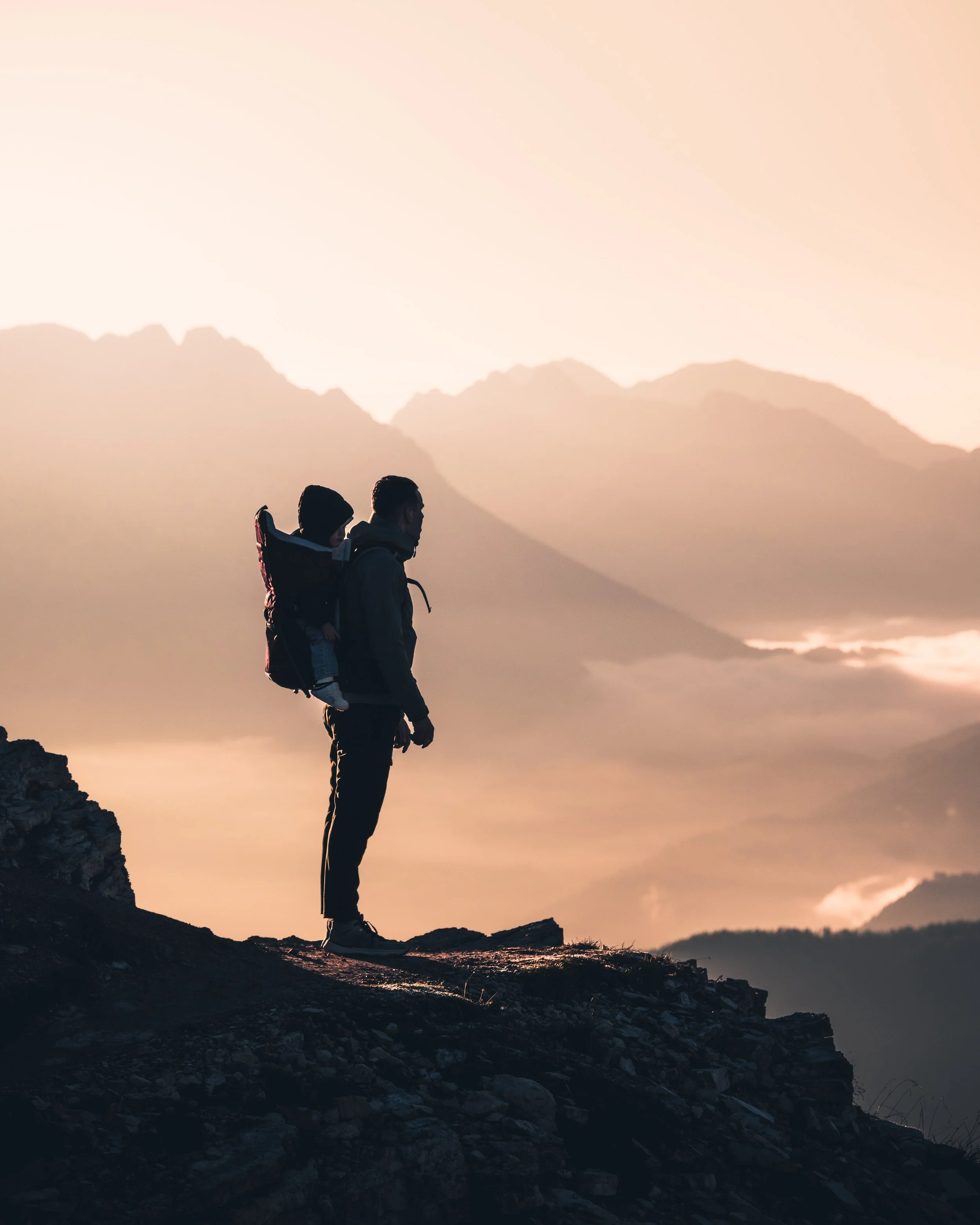 Silhouettes of two people, possibly a parent and child, standing on rocky terrain during sunset with a mountain range in the background.
