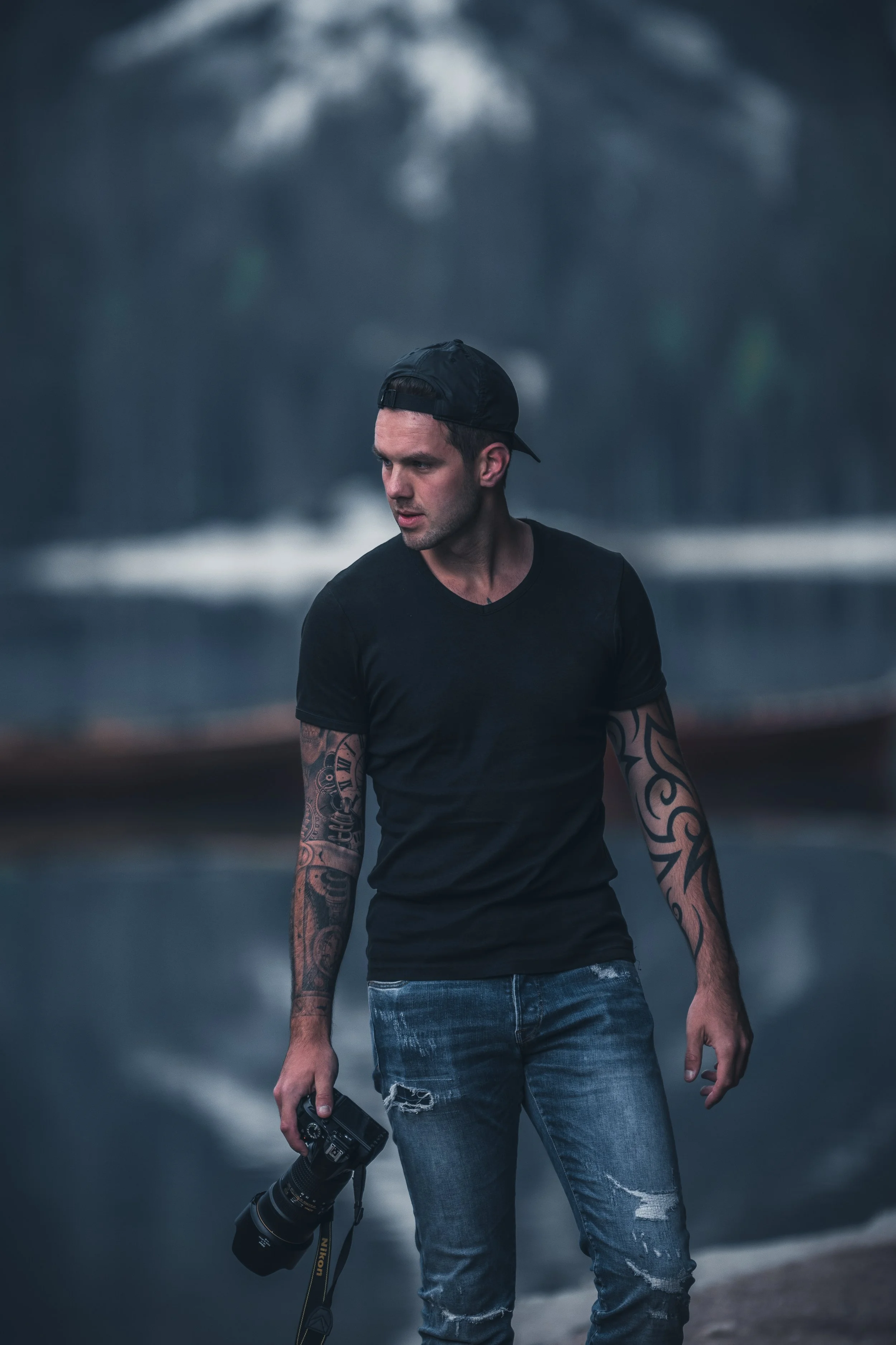 A young man with tattoos on his arms, wearing a black baseball cap backwards, a black t-shirt, and distressed jeans, holding a camera with a large telephoto lens in his right hand, standing outdoors near a road with blurred trees and cloudy sky in the background.