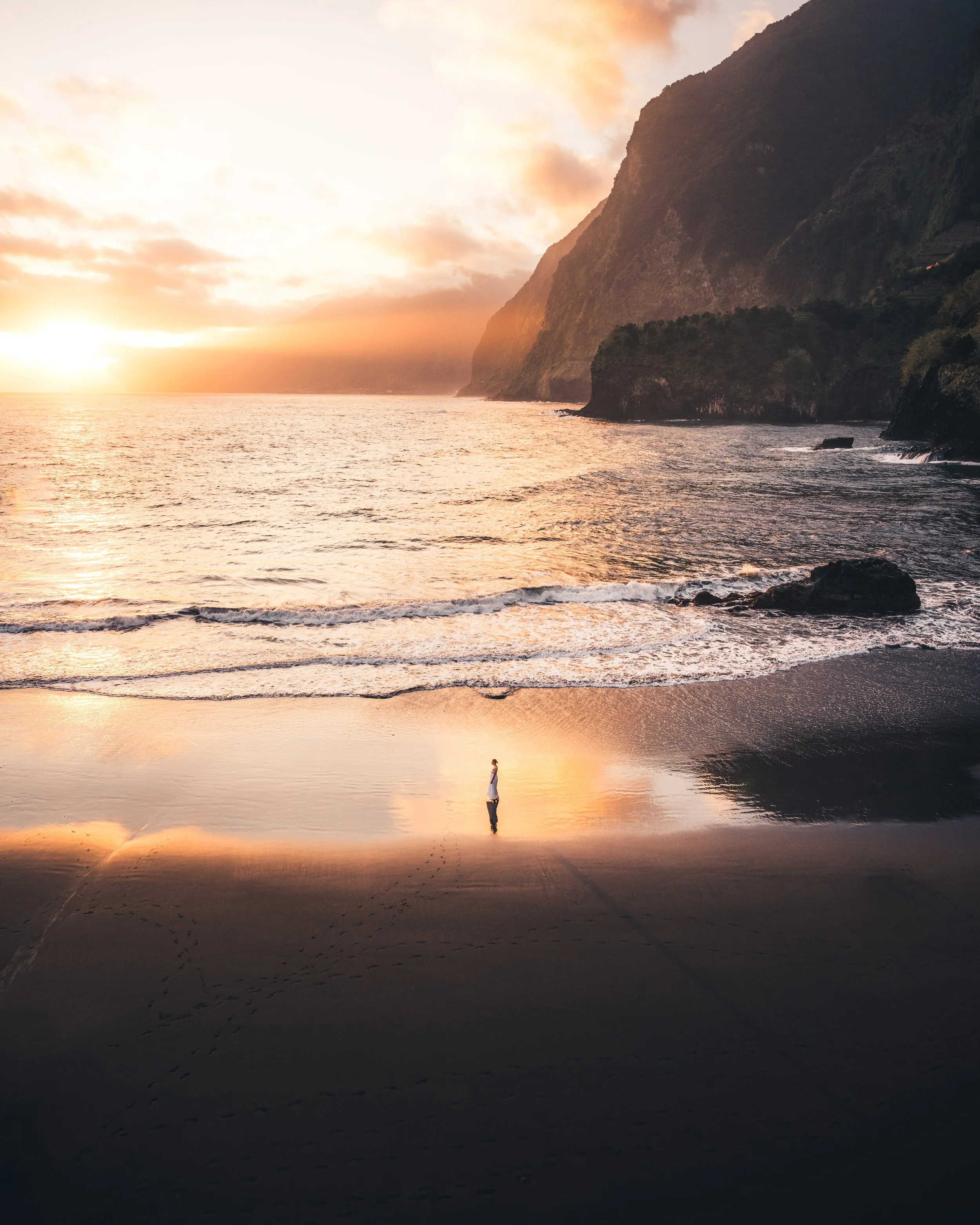 Sunset over the ocean with cliffs on the right, calm waves, and a person walking along the shore reflected in wet sand.