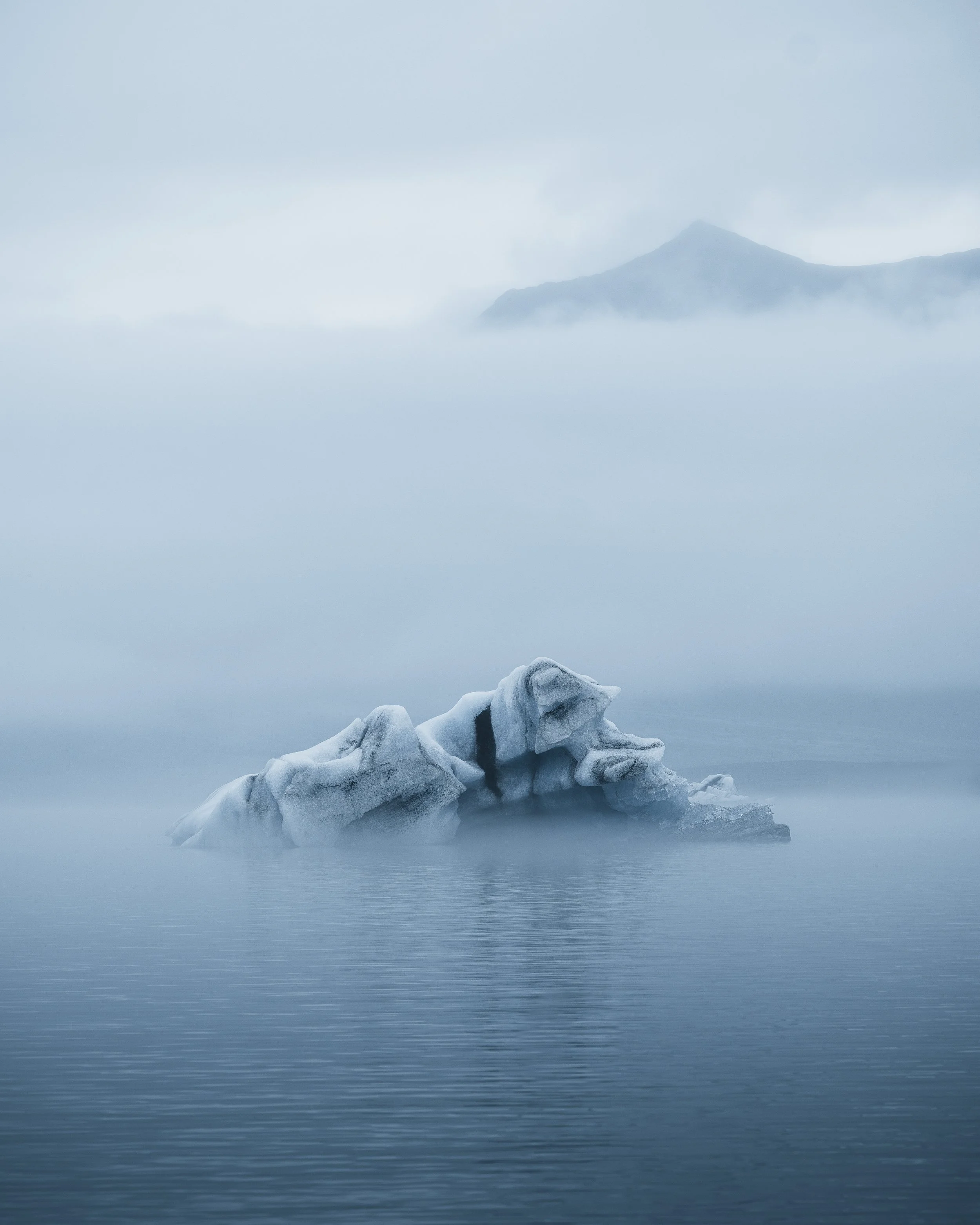 Iceberg floating in calm water with foggy mountain in the background.