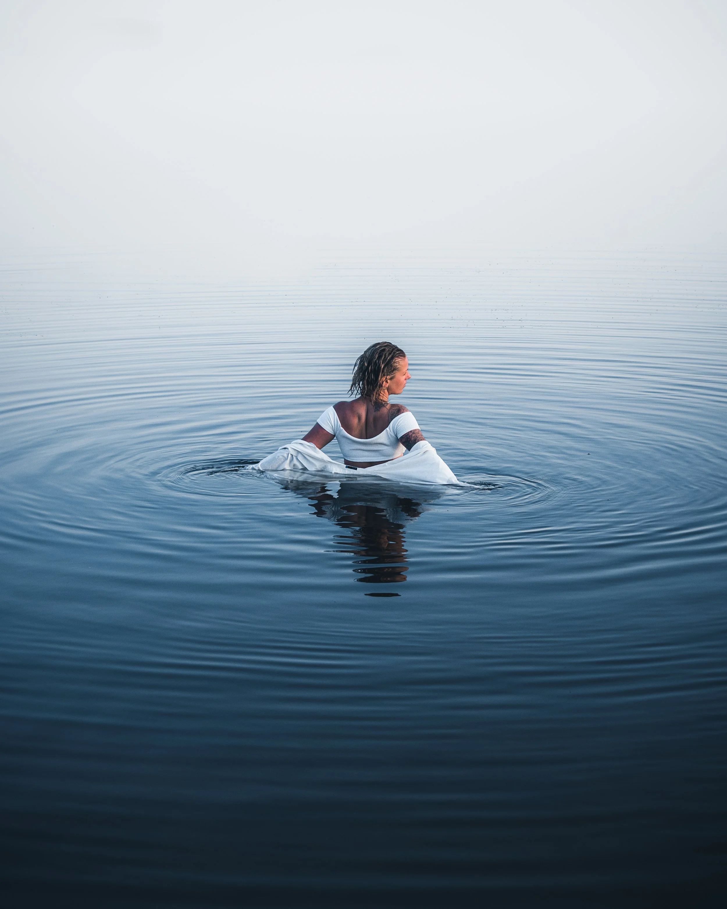 A woman with wet hair and tattoos sitting in calm water, wearing a white top and skirt, with misty, overcast sky.
