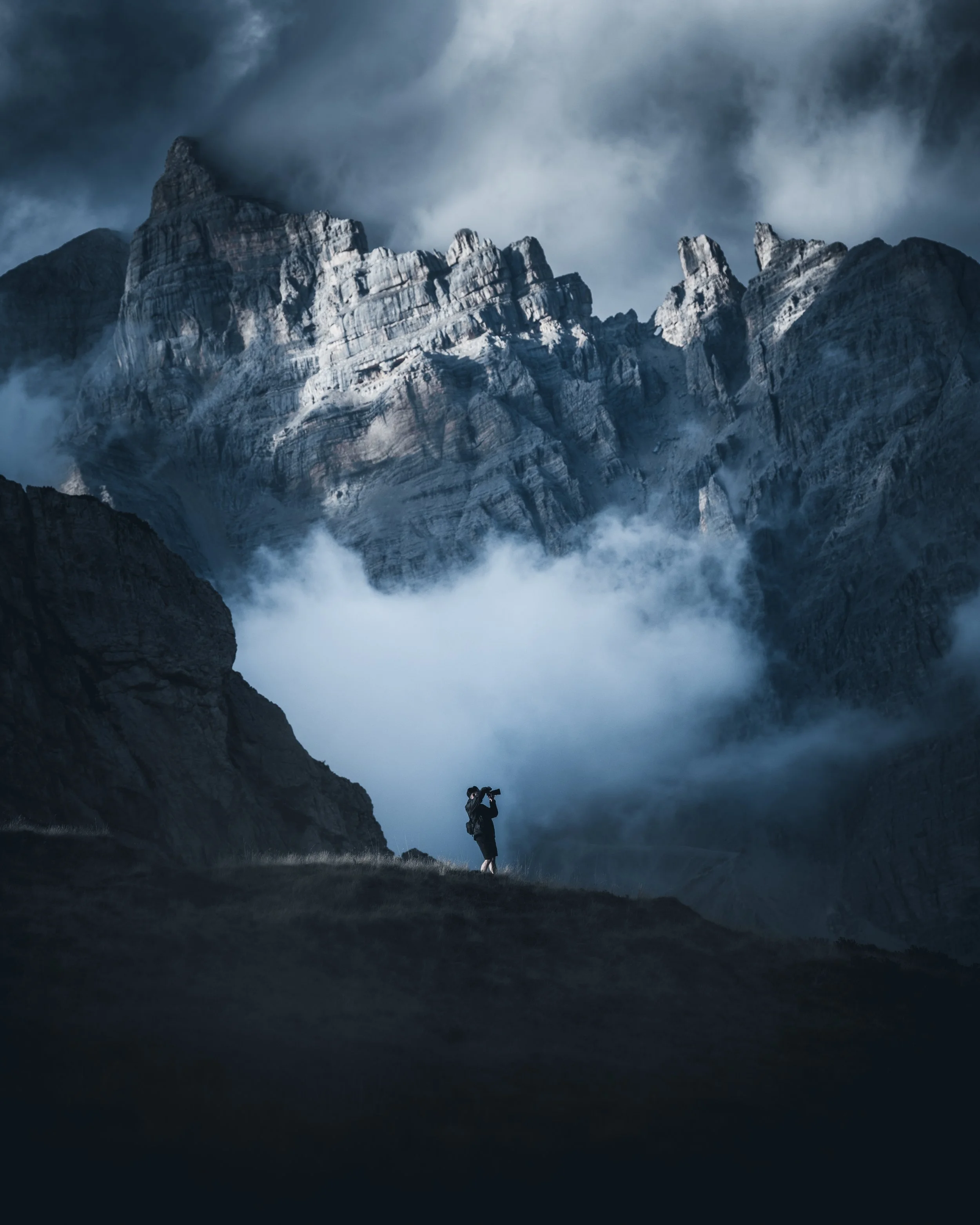 A lone hiker in dark clothing standing in a valley surrounded by tall, snow-capped mountain peaks shrouded in mist and clouds.