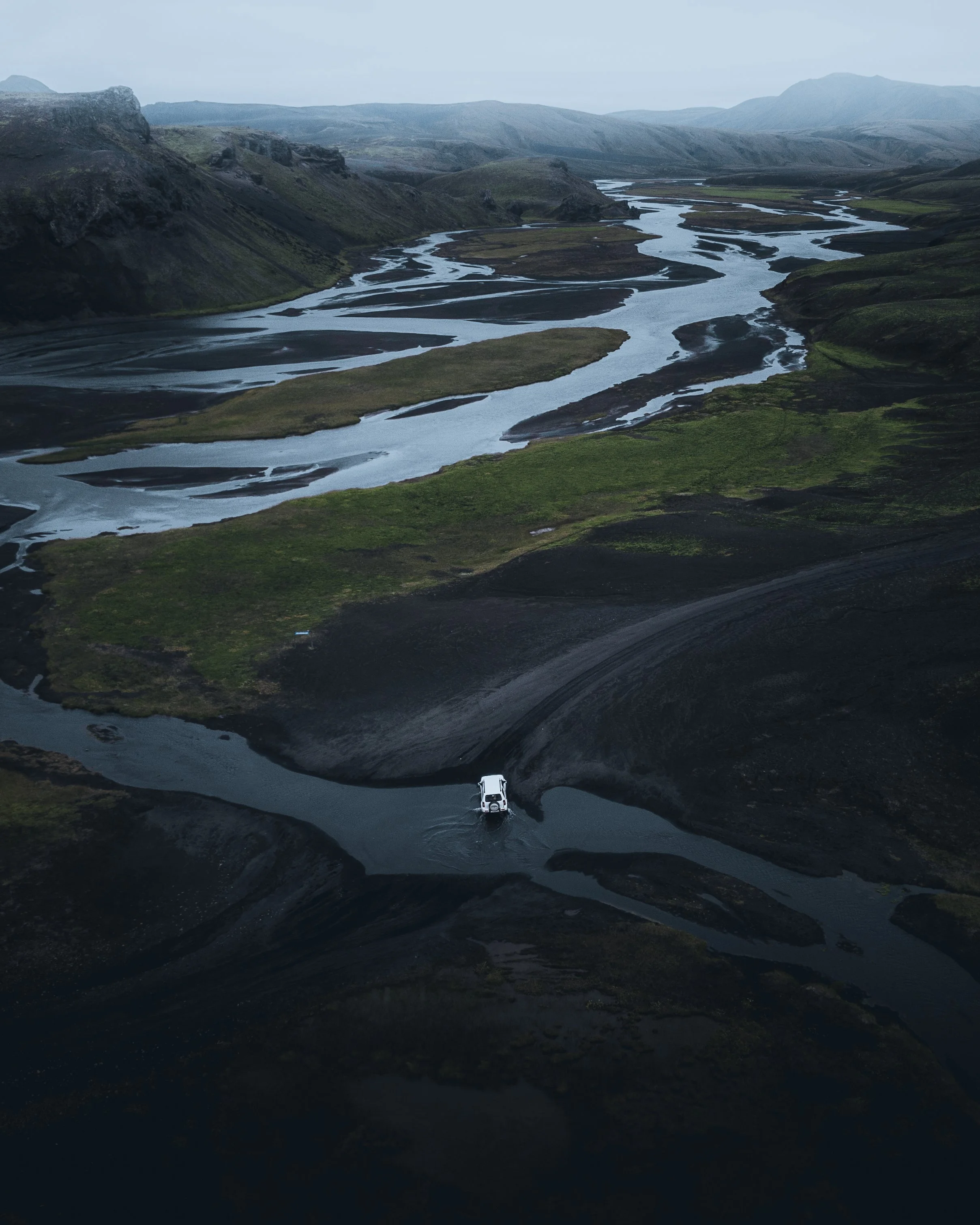 A white vehicle driving through a shallow river in a remote landscape with green hills and mountains in the background.