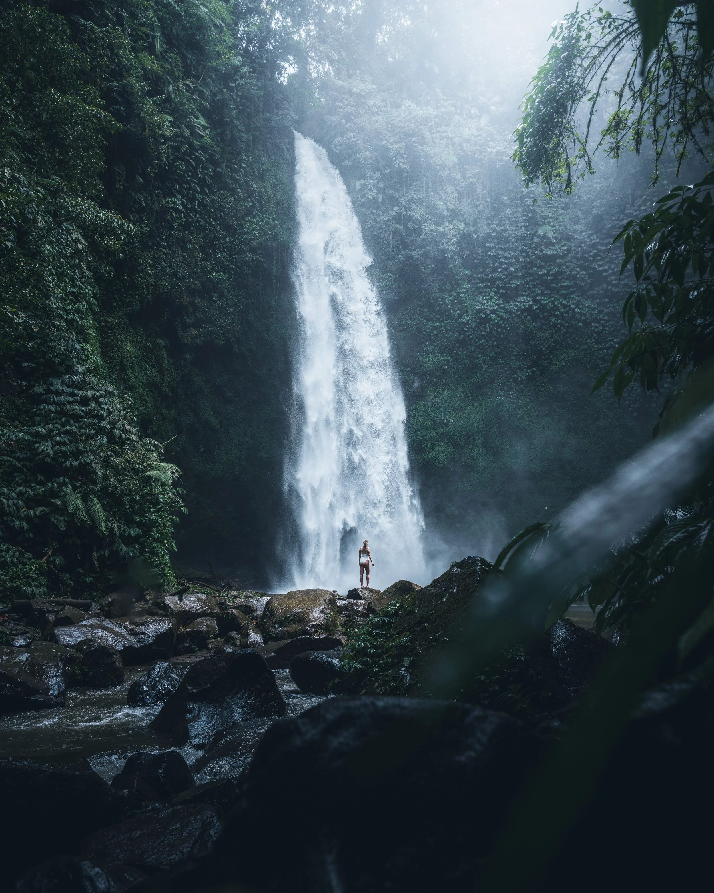 A person standing on rocks near the base of a tall waterfall in a lush, misty jungle.