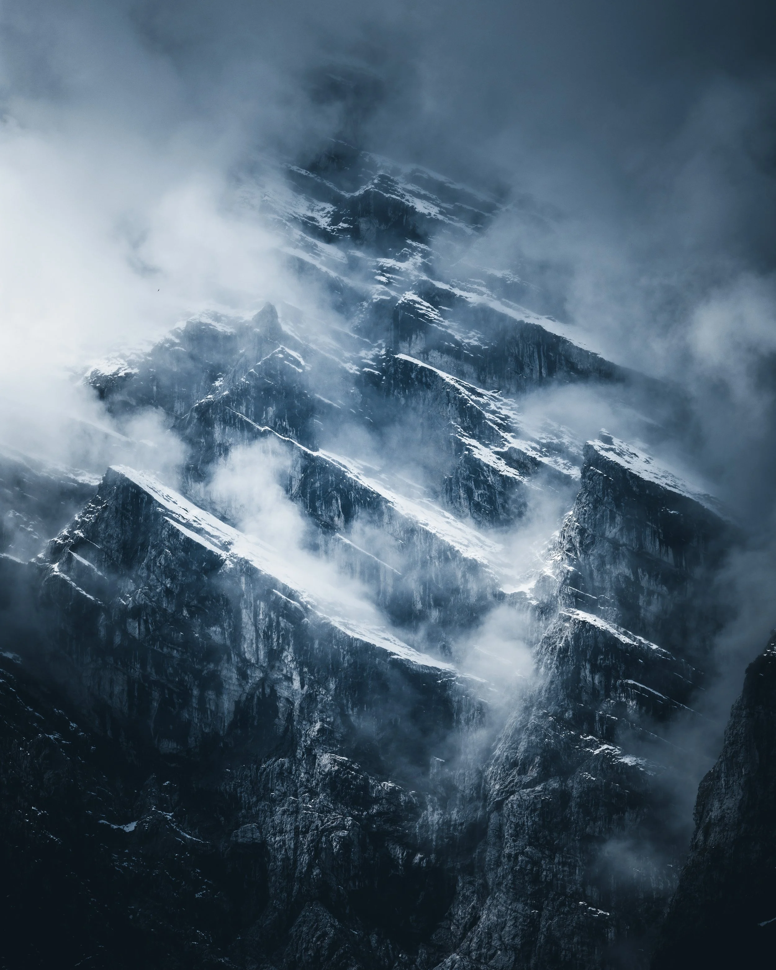 Snow-capped mountain peaks shrouded in clouds and mist, with rugged rocky slopes underneath.