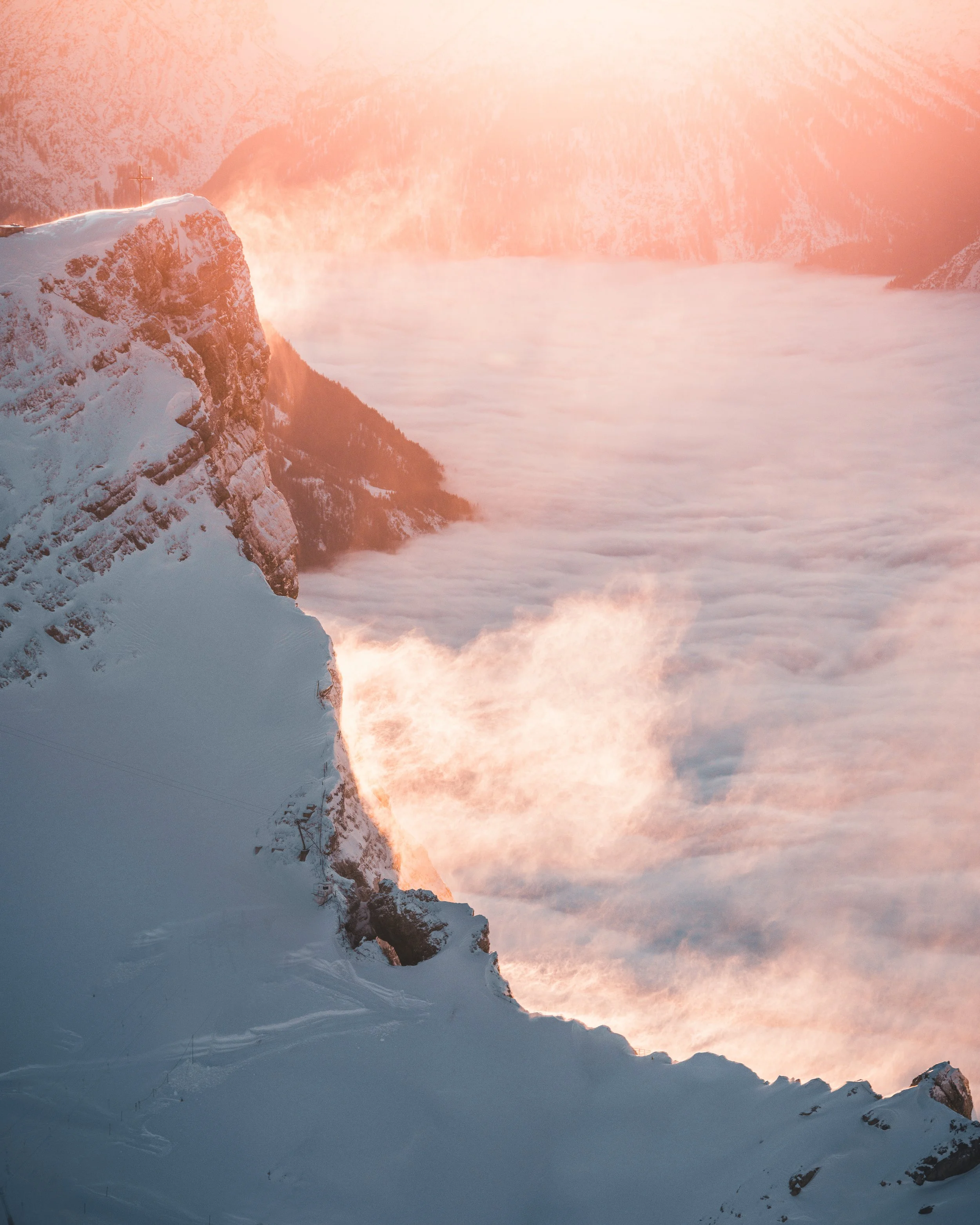 Snow-covered mountain slopes with a misty sea or lake below, illuminated by pink and orange sunrise or sunset light.