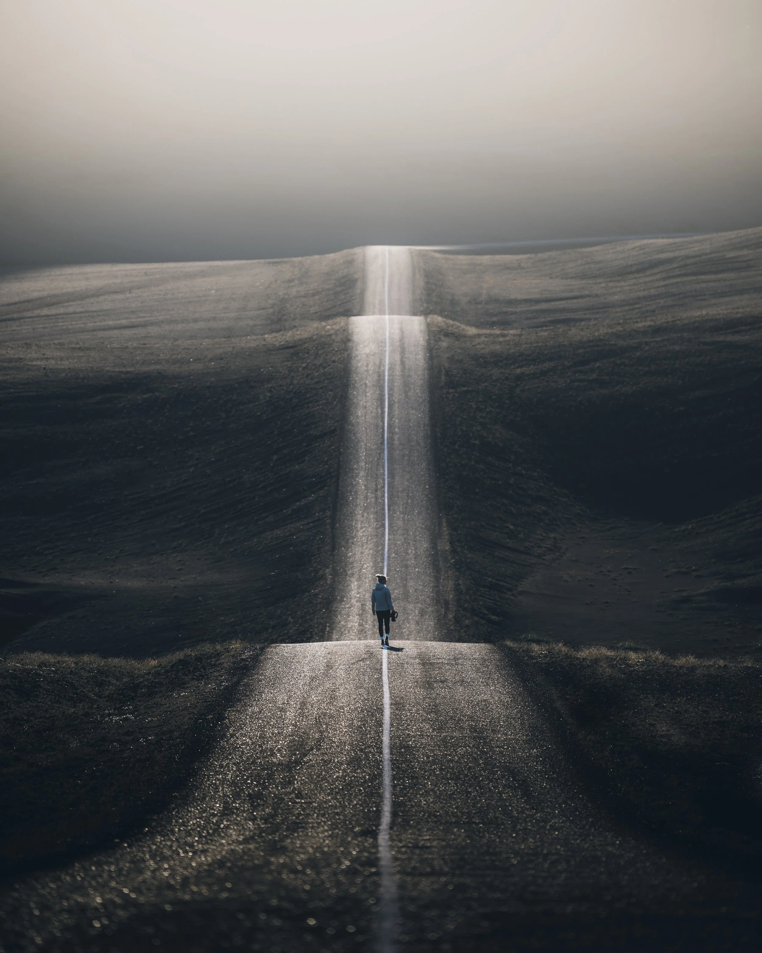 A solitary person walking down a road in a vast, empty landscape with dark and light contrasting colors, and a large, confusing mountain-like formation in the background with a long, straight light streak crossing the scene.