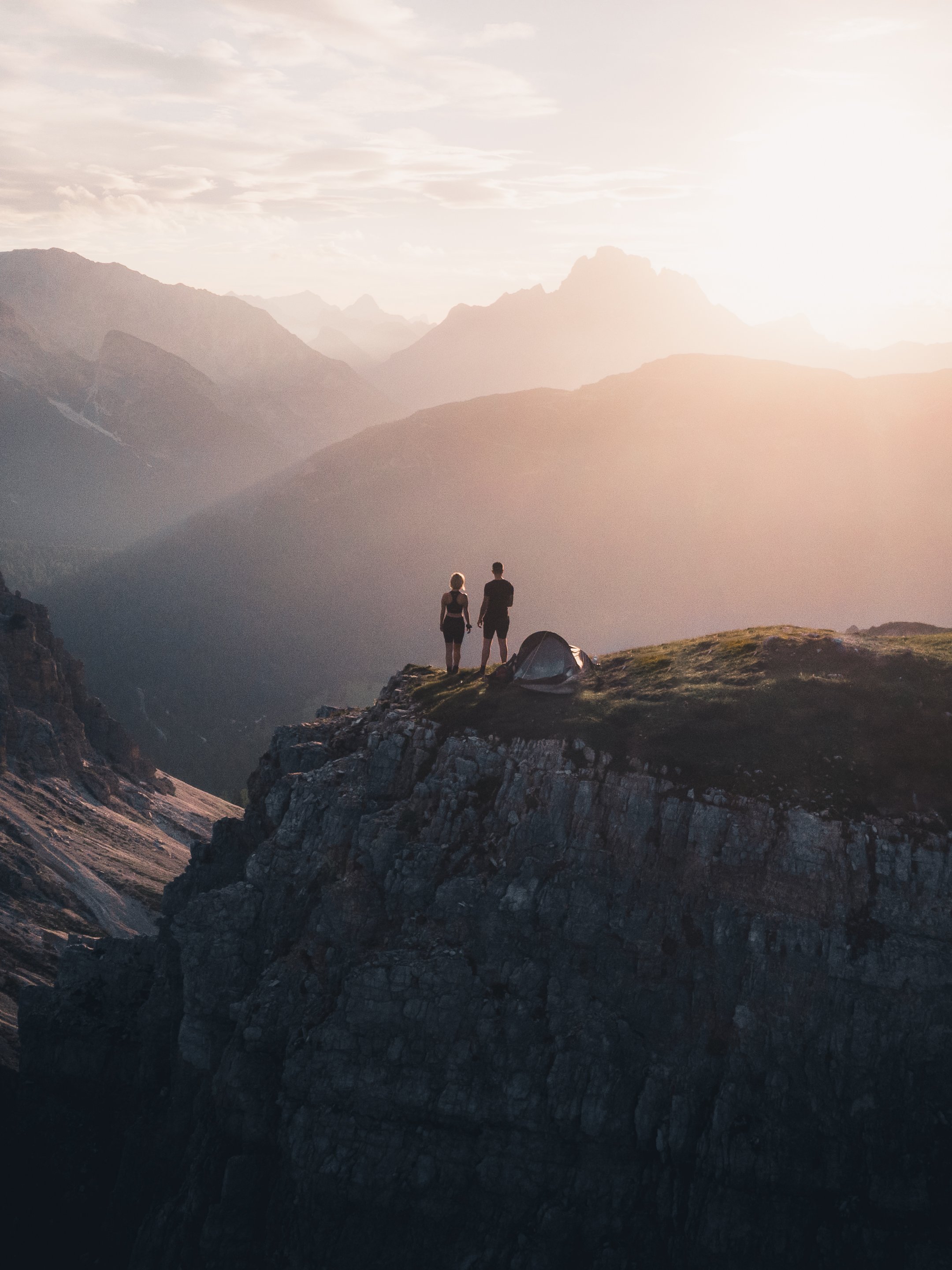 A man and woman holding hands standing on a rocky ledge, facing a mountain range during sunset or sunrise.