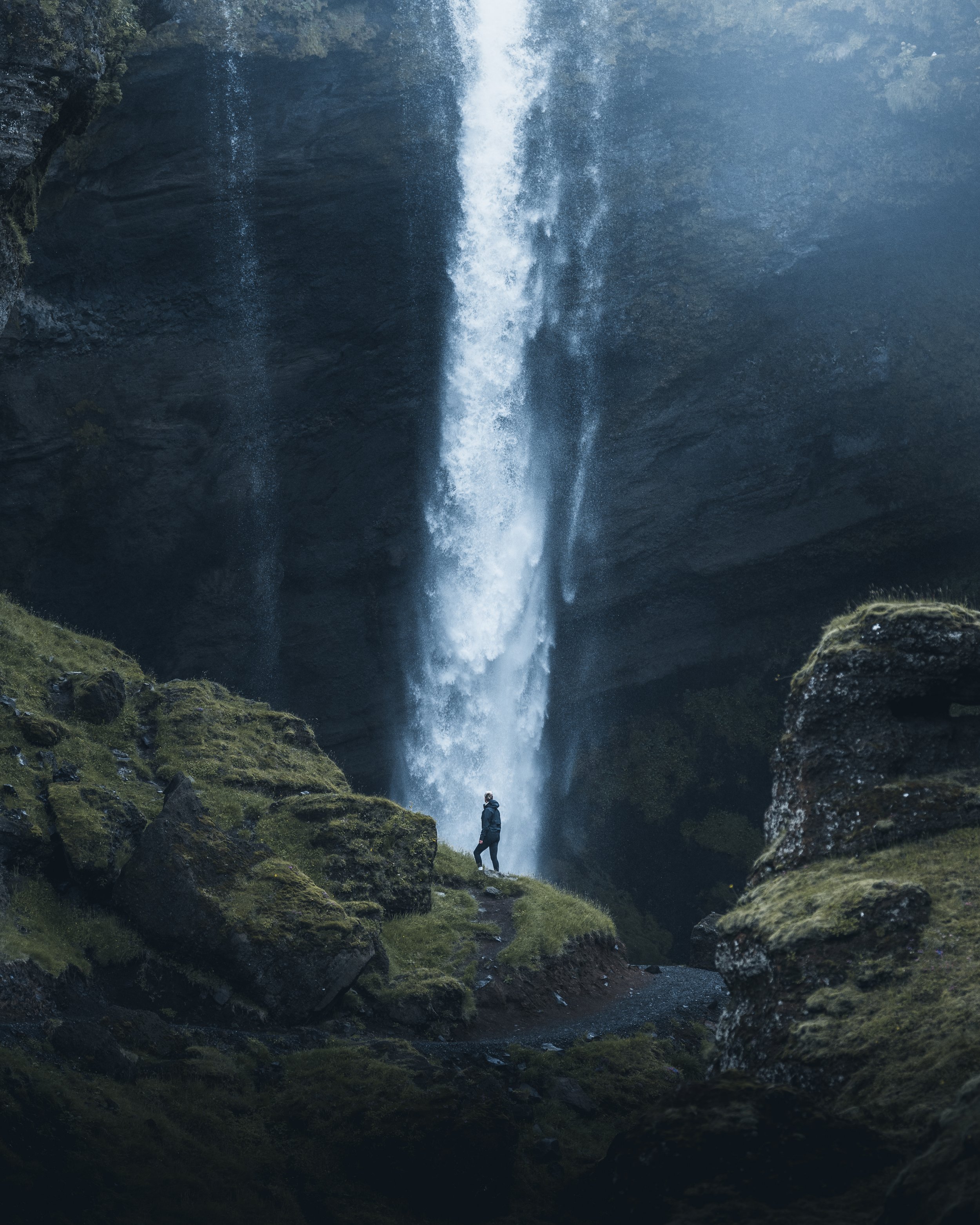 A person standing on a mossy rock during daytime, with a large waterfall cascading down a rocky cliff in the background.