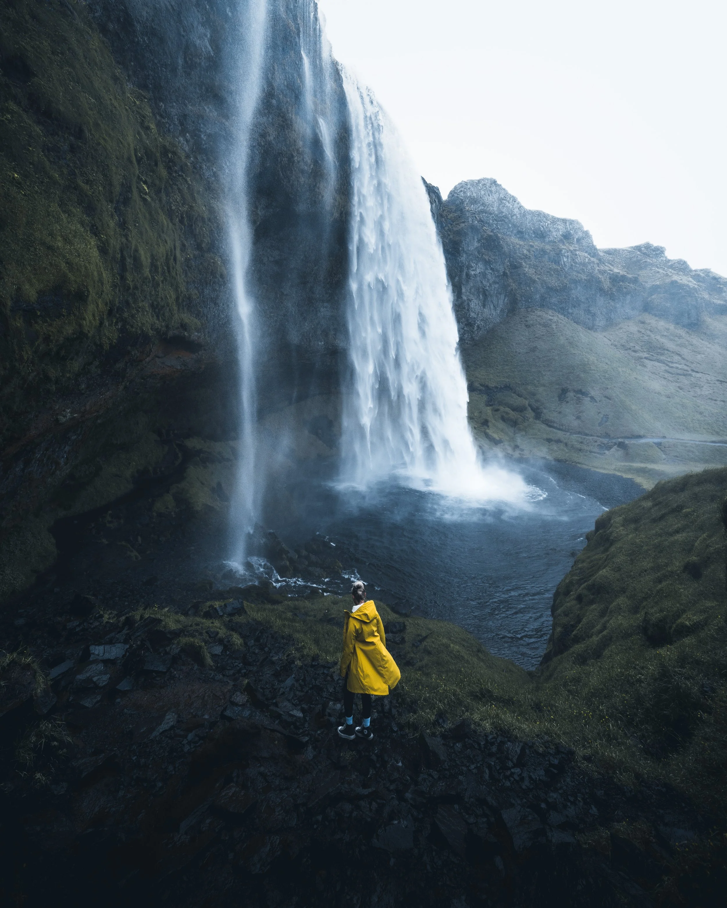 A person wearing a yellow jacket and black pants standing on rocky terrain, viewing a tall waterfall cascading down a cliff surrounded by green moss and rocks.