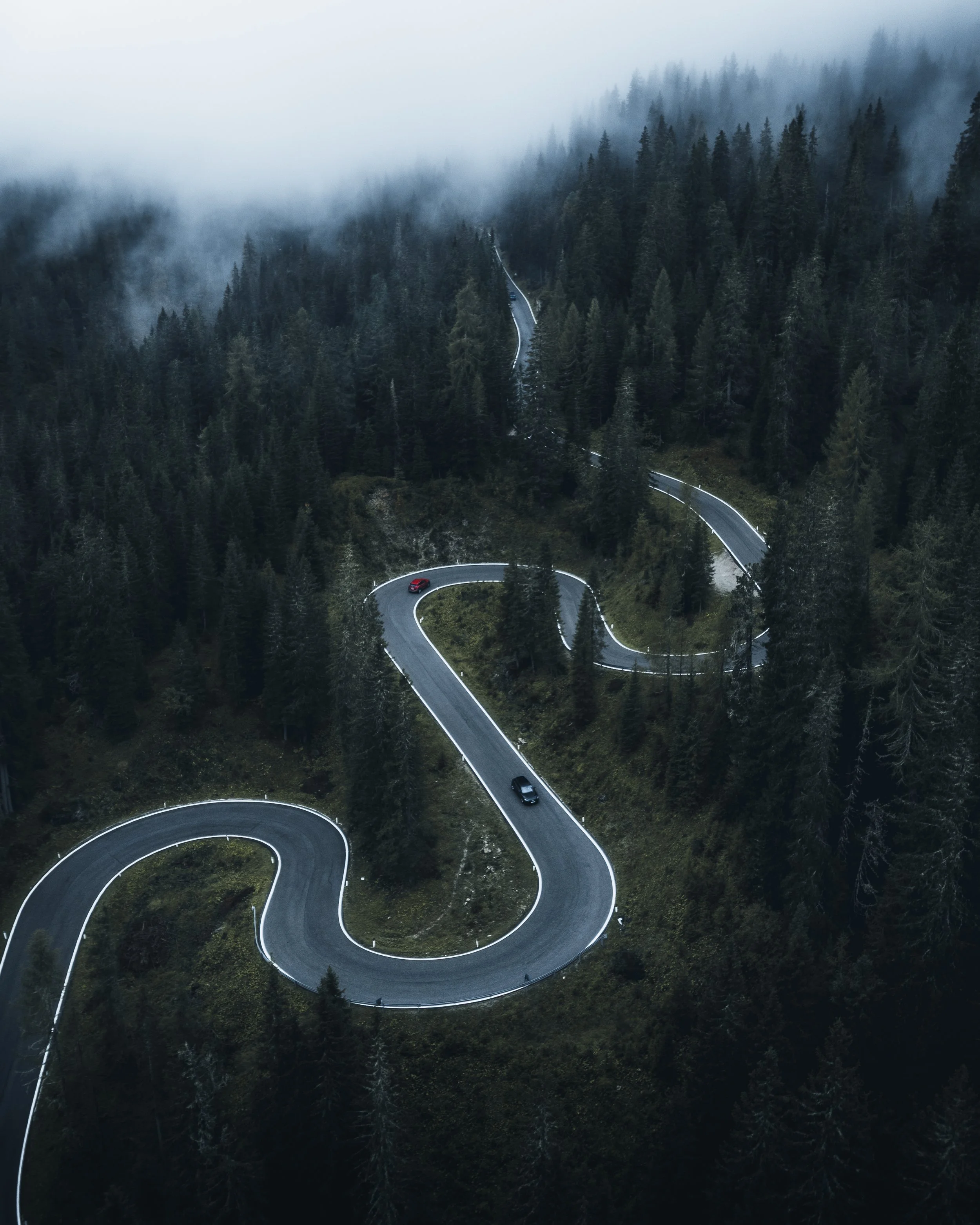 A winding mountain road through a forest, with fog and mist hanging in the air, and three cars visible on the twists and turns of the road.