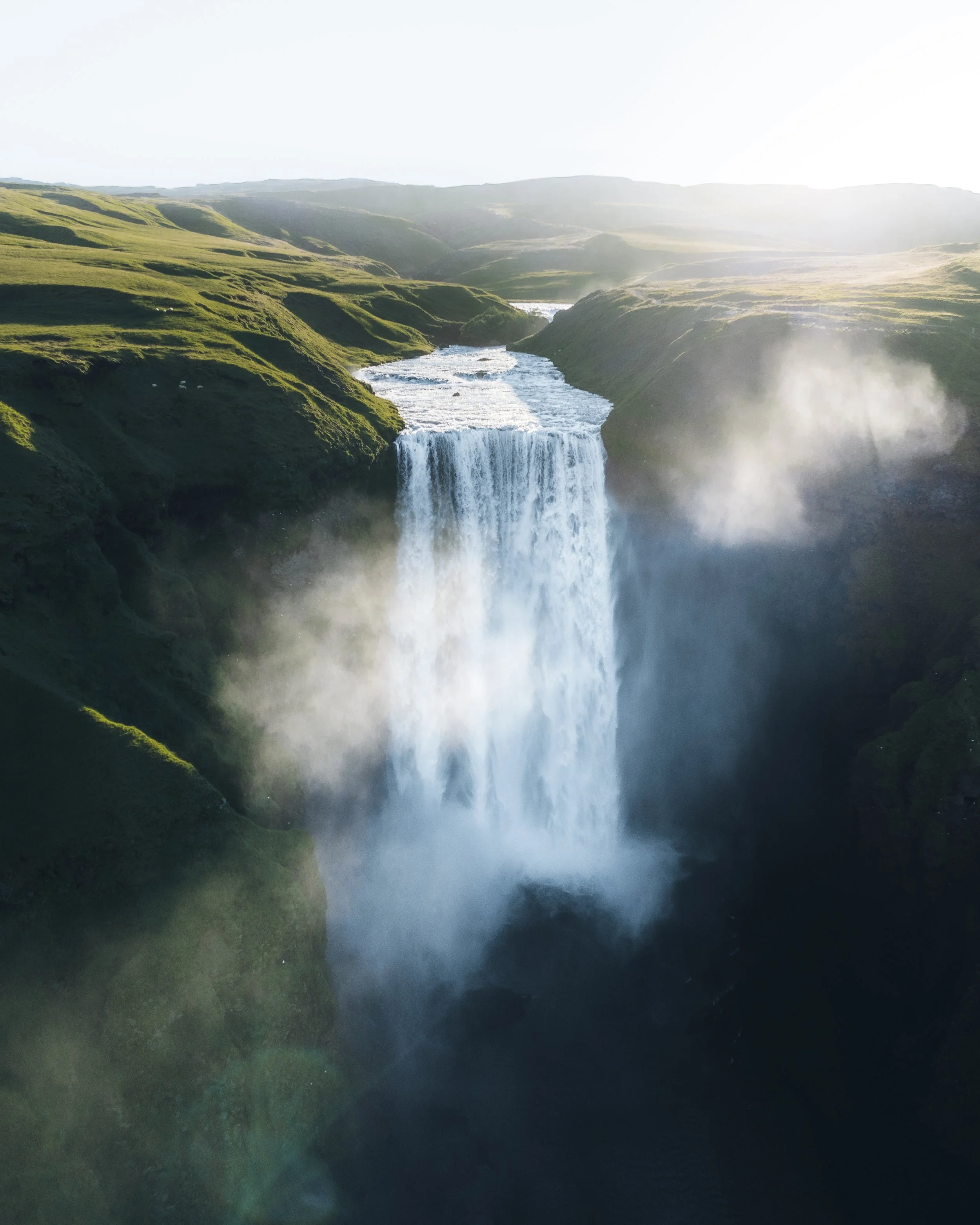 A tall waterfall cascading into a deep pool surrounded by lush green hills under sunlight.