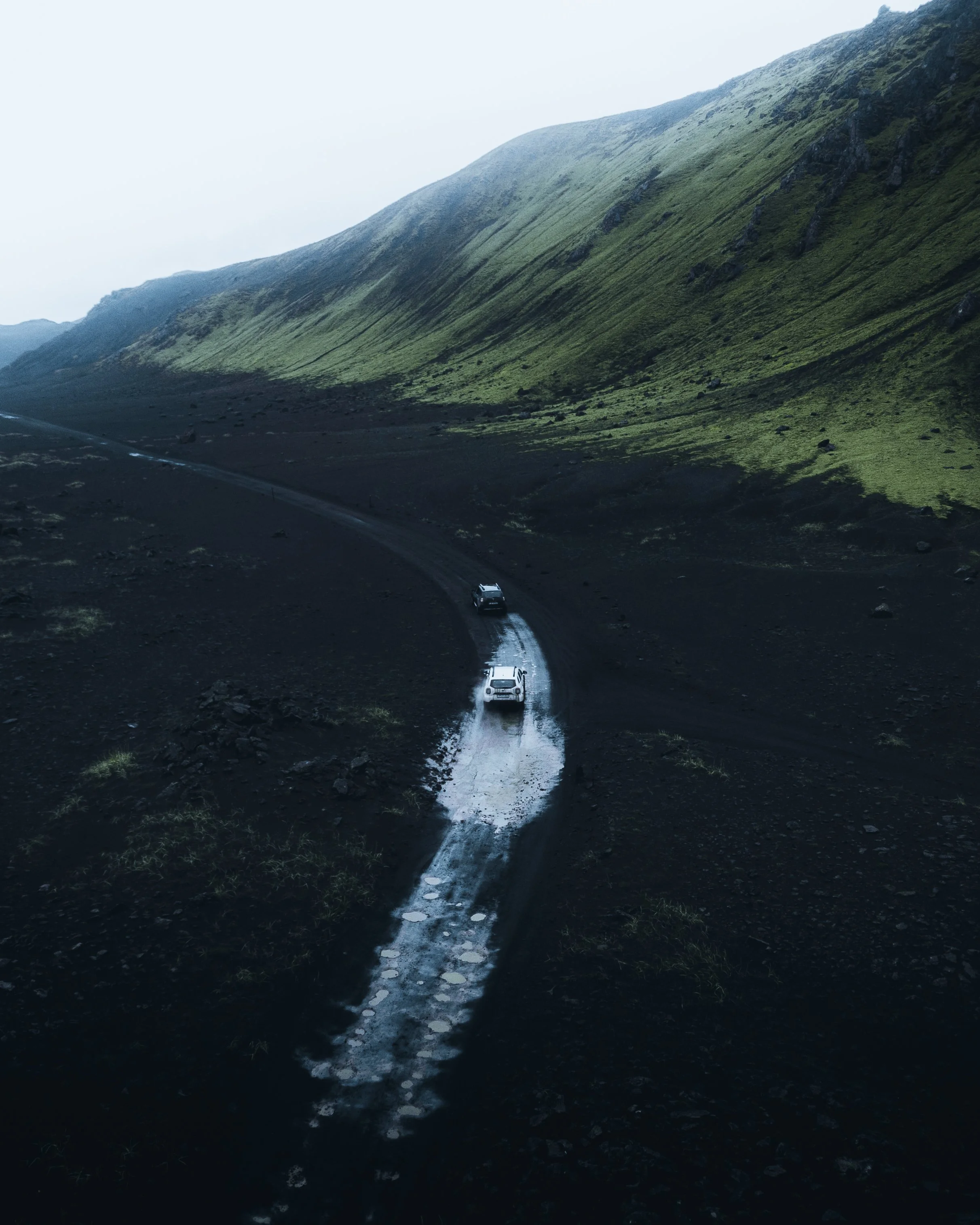 A winding mountain road with a few cars driving through a dark, wet terrain surrounded by grassy hills and rocky slopes under a cloudy sky.