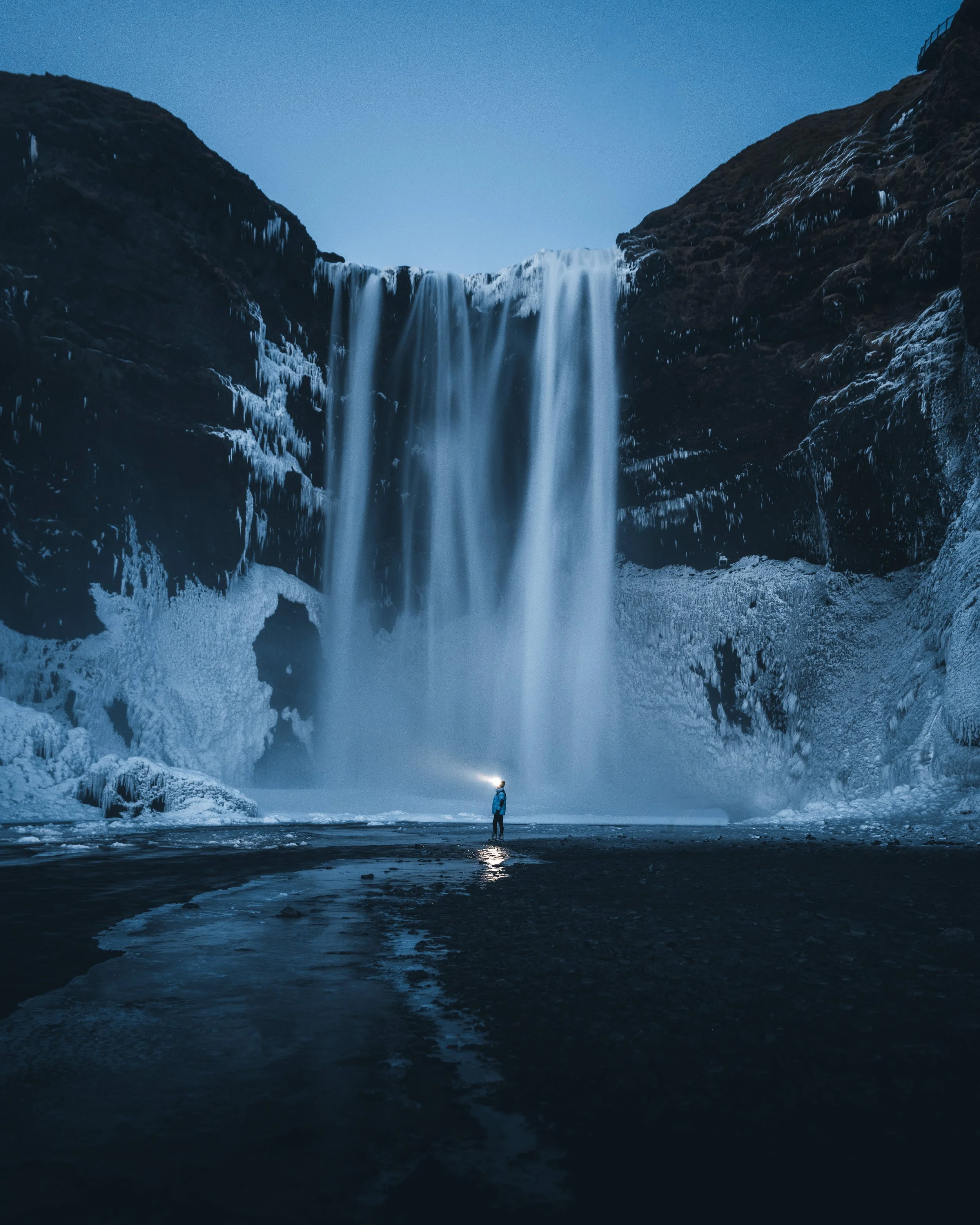 Nighttime scene of a person standing near the base of a tall, partially frozen waterfall with icy cliffs on either side, under a clear sky.