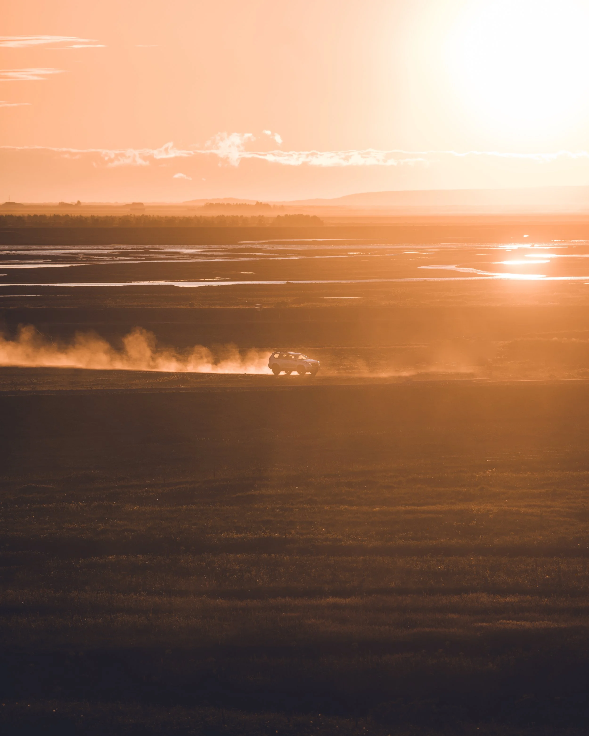 A vehicle driving on a dirt road during sunset or sunrise with water reflections, open flat landscape, and orange sky.