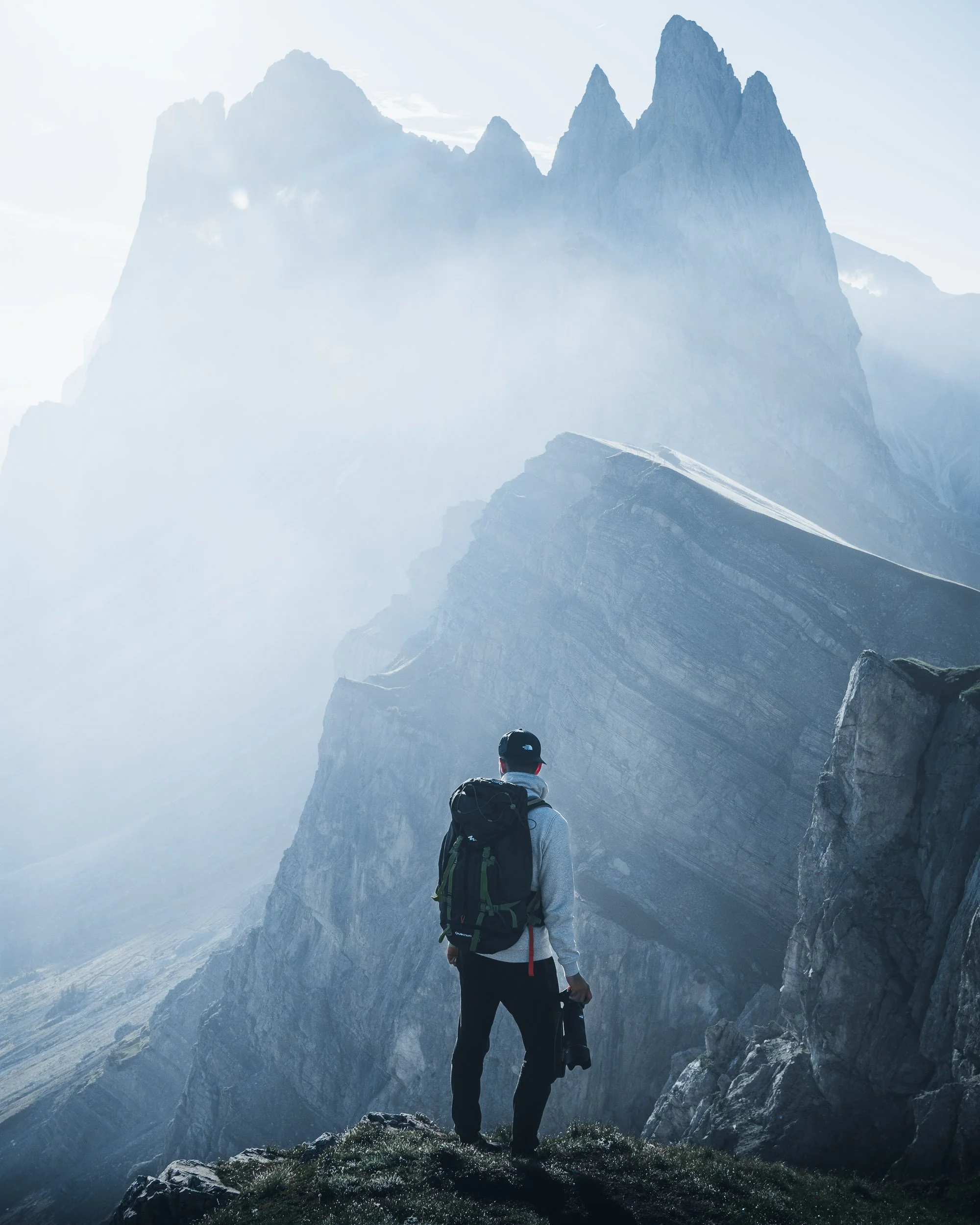 A person wearing a cap, grey jacket, and carrying a backpack stands on a rocky ledge in a mountain landscape, looking at steep, jagged peaks shrouded in mist.