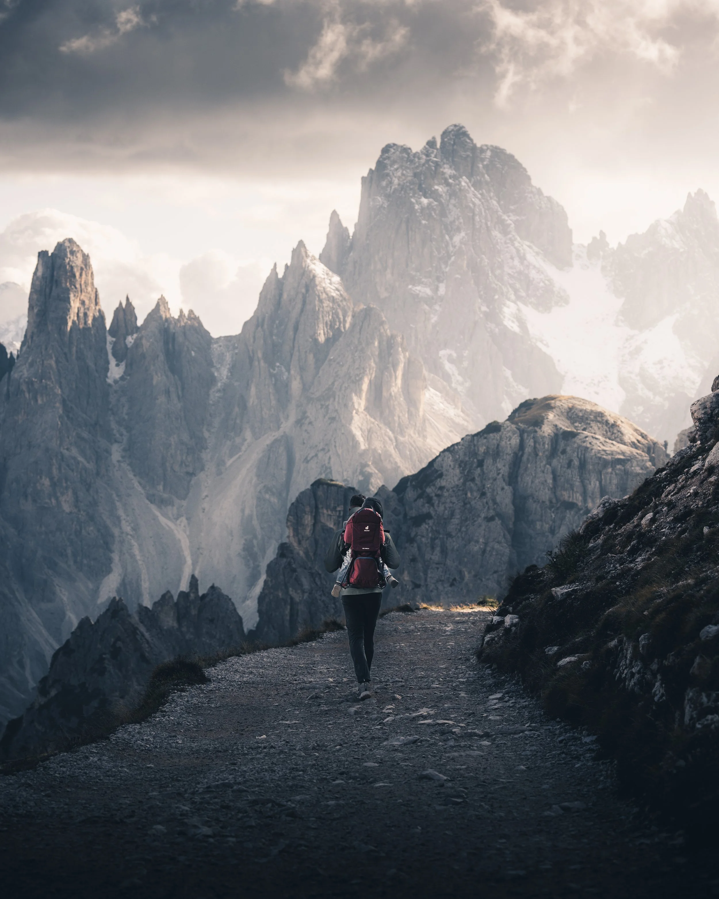 A person with a pink backpack hiking on a rocky trail in a mountainous area with snow-capped peaks and dark clouds in the sky.