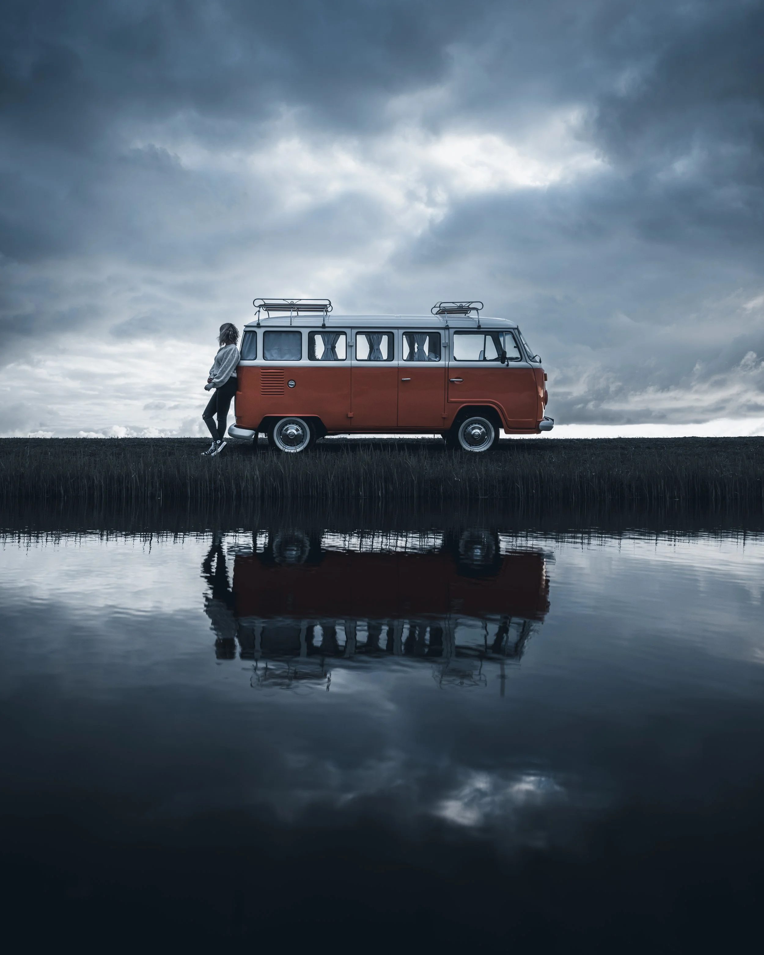 A person leaning against a vintage red and white van parked beside a calm body of water, under a cloudy sky.