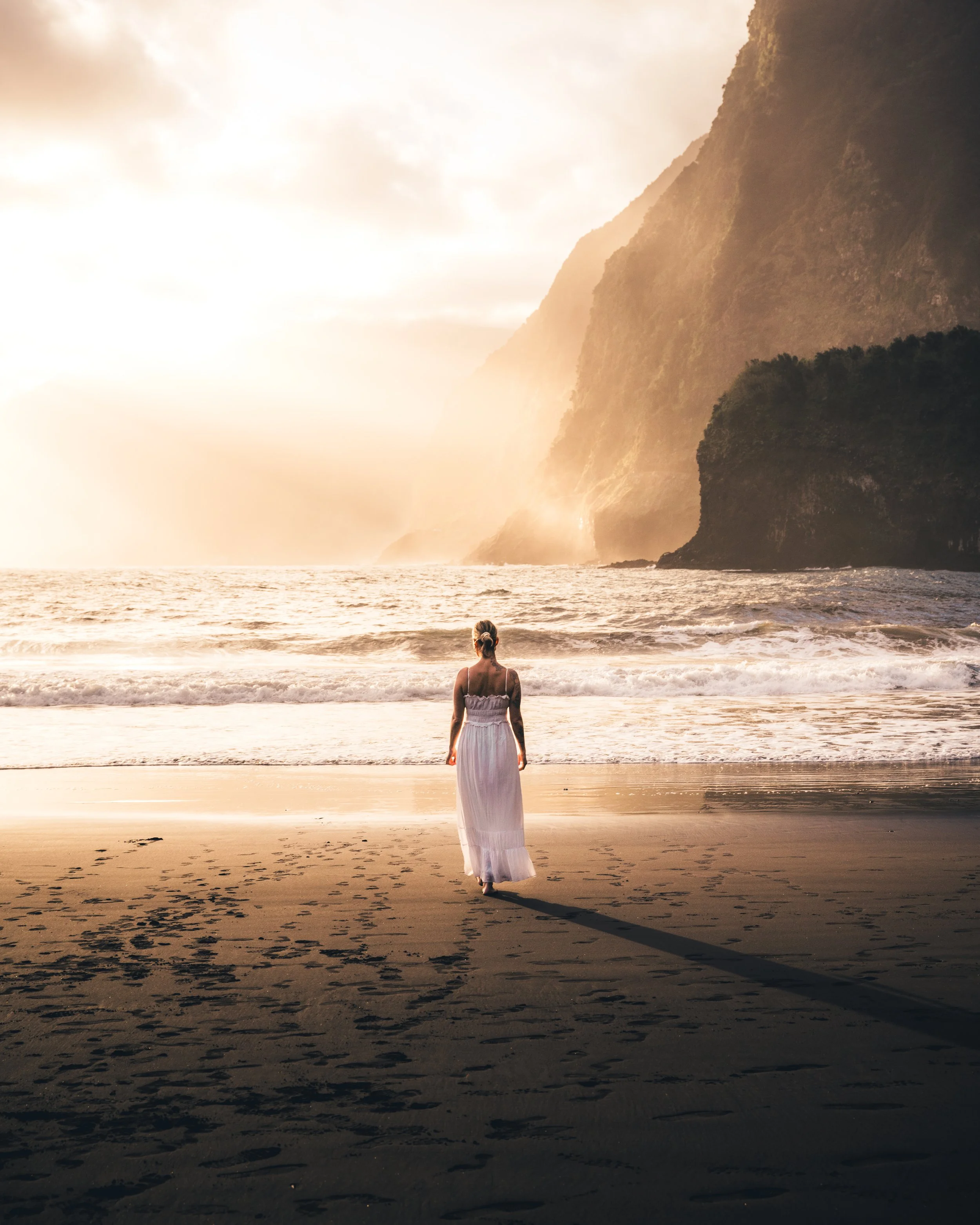 A woman in a white dress standing on the beach facing the ocean, with mountains and a cloudy sky in the background during sunset.