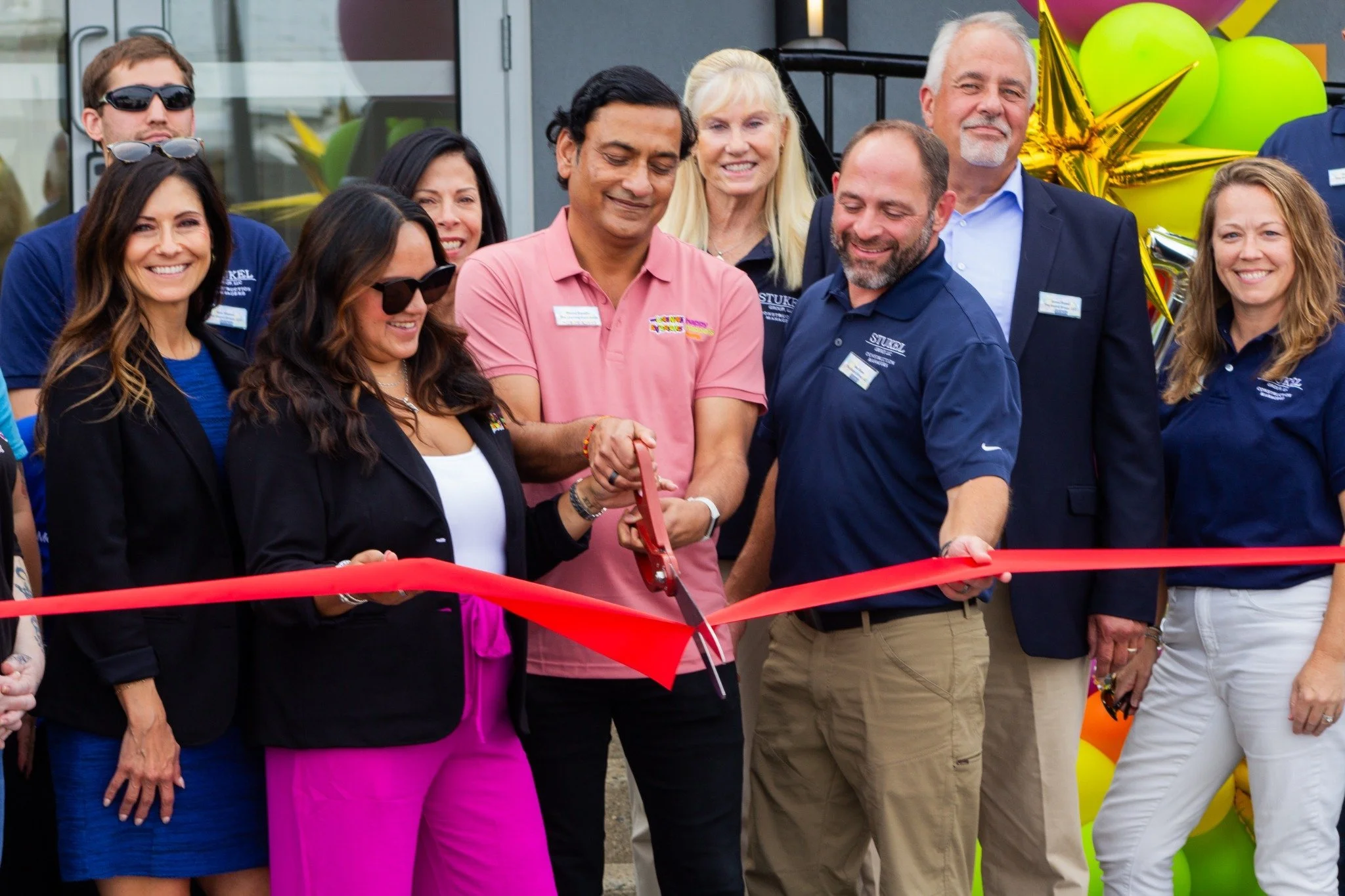 People gathered for a ribbon-cutting ceremony, with some holding scissors, in front of balloons and decorations. The group is smiling and dressed in business casual attire.