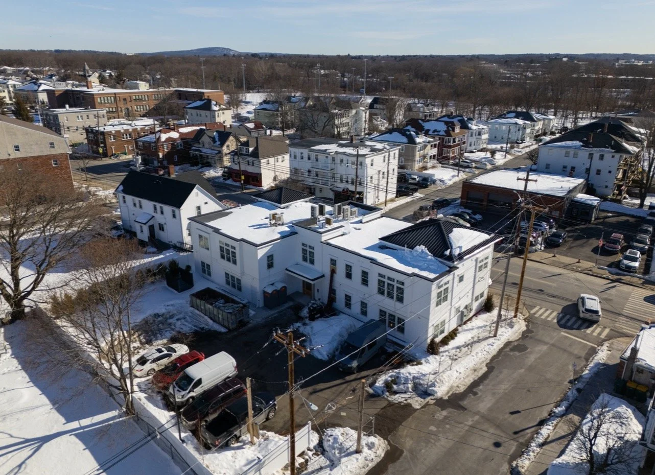 An aerial view of a snowy residential neighborhood with multi-story houses, parking lots, and streets, under a clear blue sky.