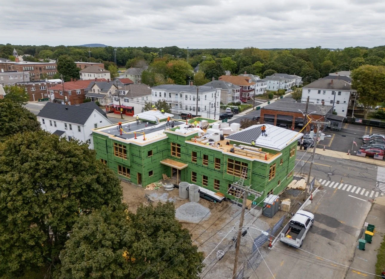 Aerial view of a multi-story building under construction with green exterior sheathing, workers on the roof, and surrounding residential neighborhood with trees and parked cars.