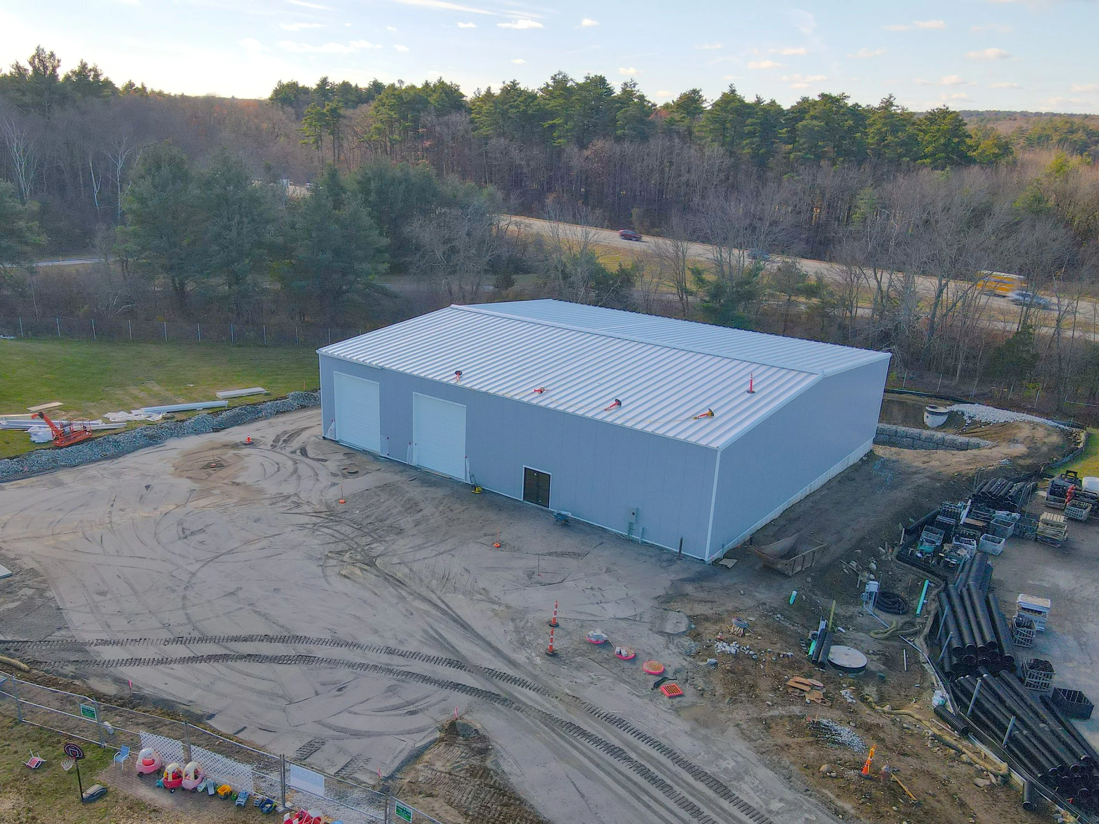 An aerial view of a construction site with a large white industrial building, dirt roads, construction equipment, and storage pipes, surrounded by trees and a highway with passing cars.