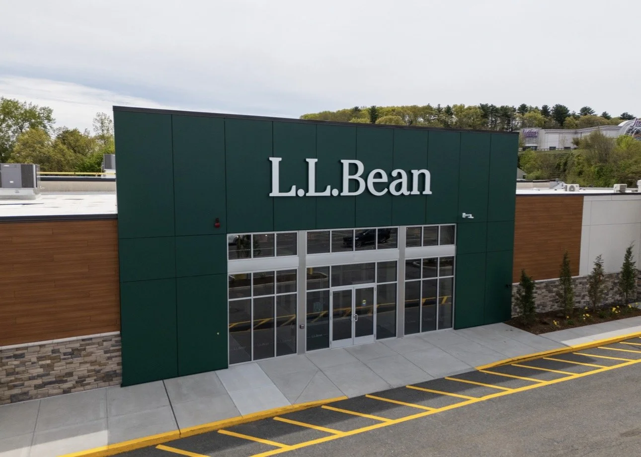 L.L.Bean retail store exterior with green and wood facade, large glass doors, parking lot with yellow striped spaces, and a background of trees and a cloudy sky.