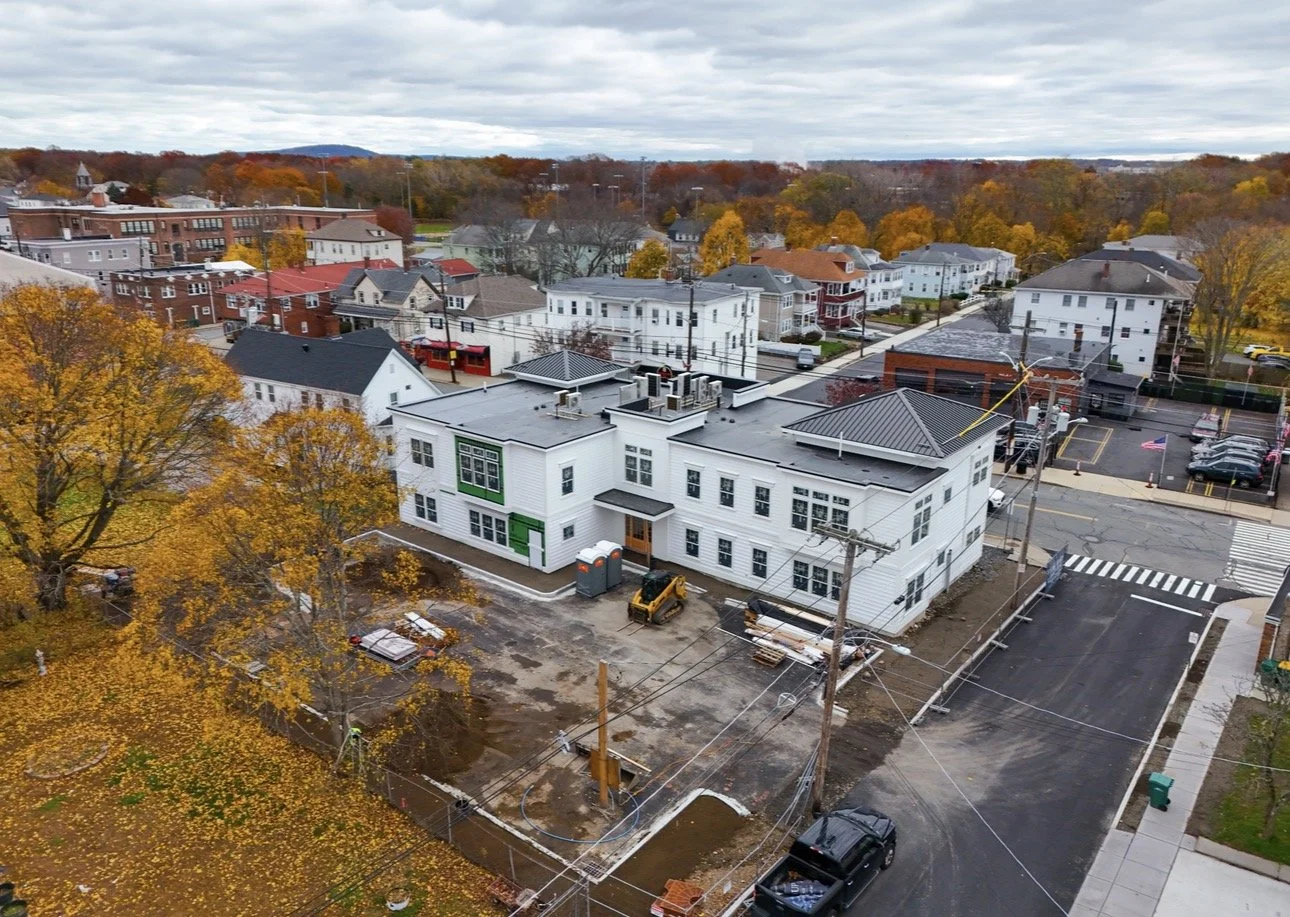 Aerial view of a construction site with a large white building, surrounded by a partially paved driveway and trees with fall foliage, in a neighborhood with additional residential and commercial buildings under a cloudy sky.