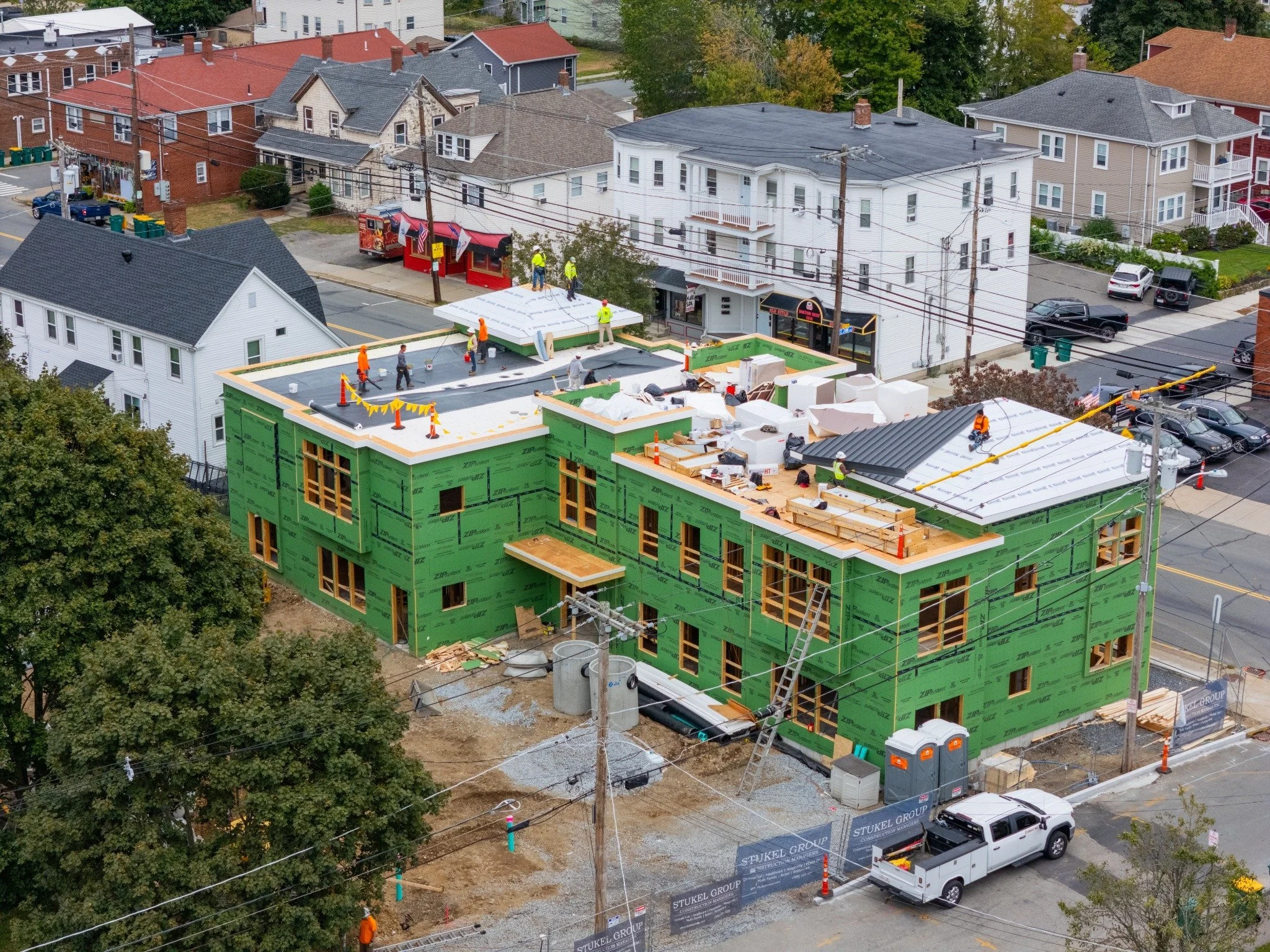 Construction workers building a multi-story residential building with green sheathing, working on the roof and walls, with various tools and materials around. Aerial marketing photo of a waterfront property in Rhode Island designed for real estate ca