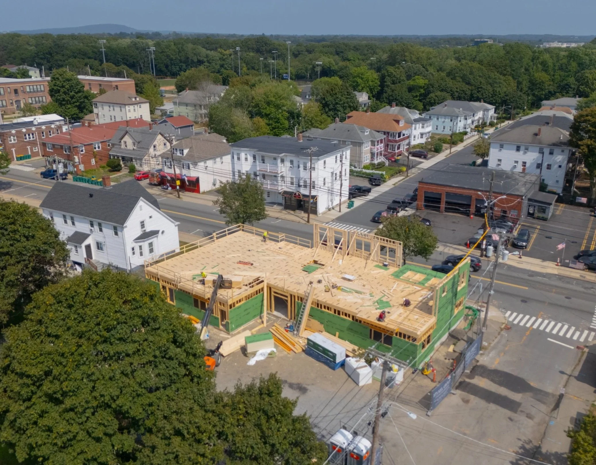 Aerial view of a construction site with wooden framing in a residential neighborhood, surrounded by trees and streets with parked cars.