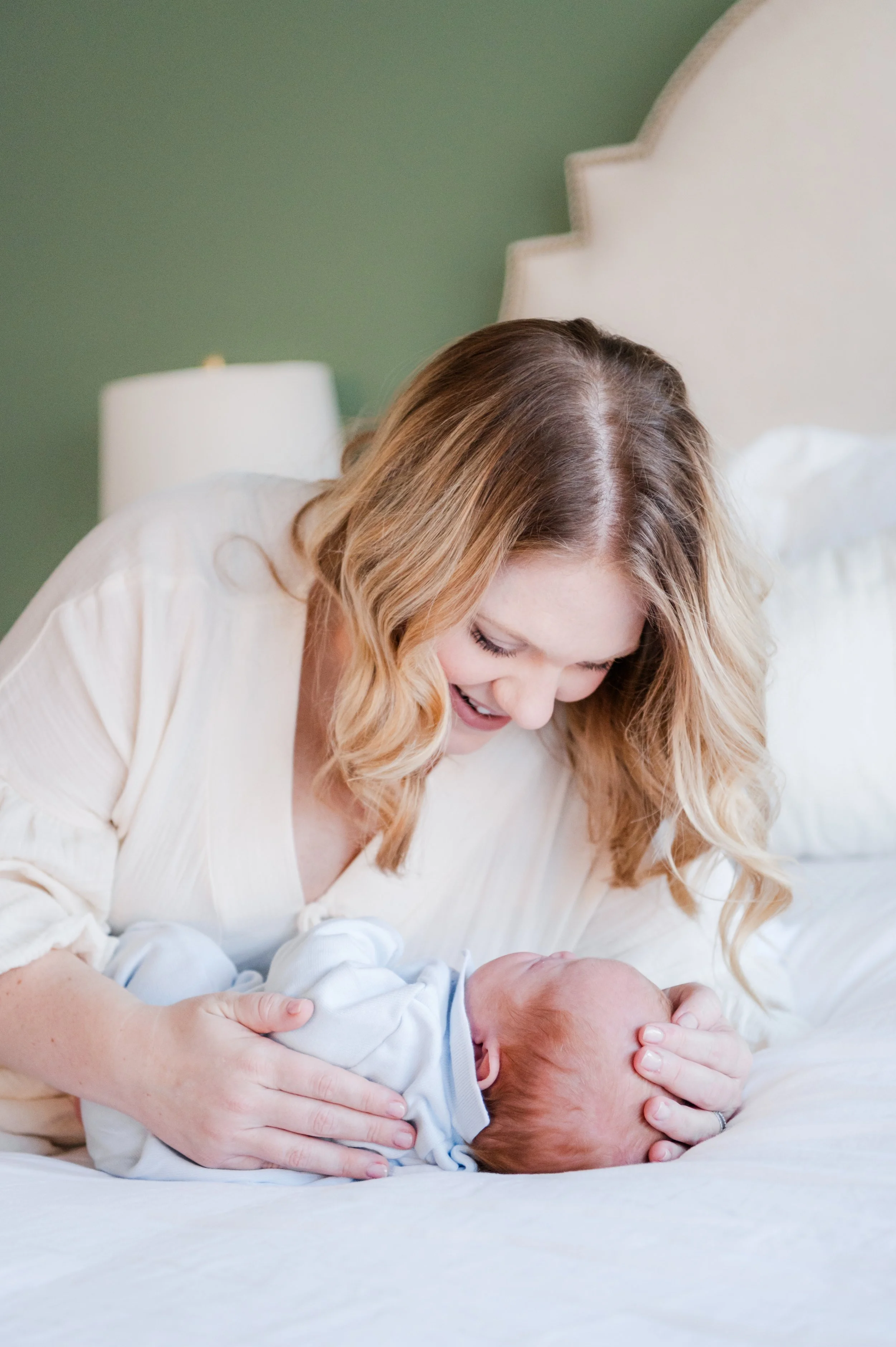 A woman with blonde wavy hair smiling and holding a newborn baby on a bed with white bedding and a green wall in the background.