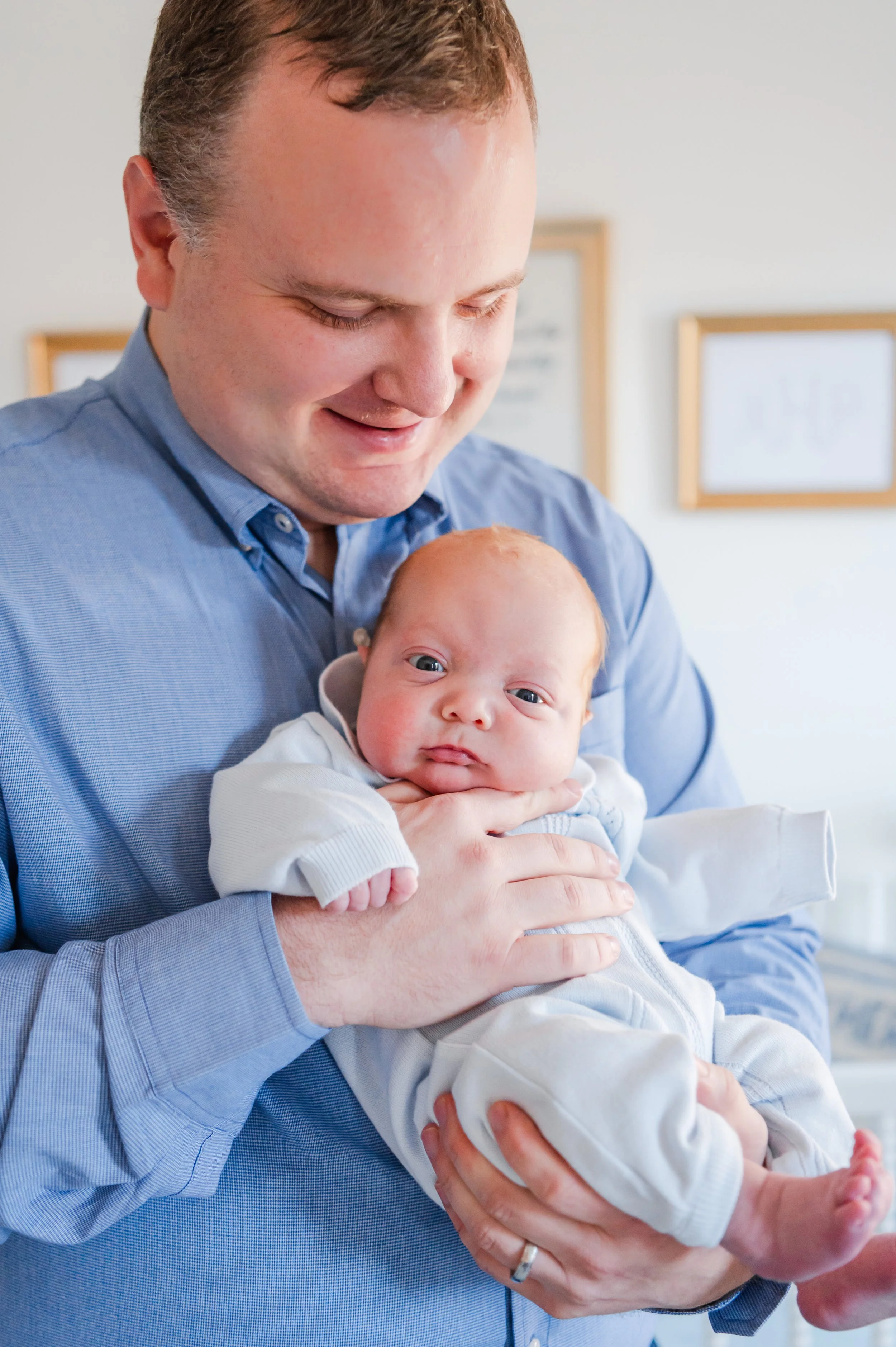 A man in a blue shirt holding a baby in a white outfit indoors with framed wall art in the background.