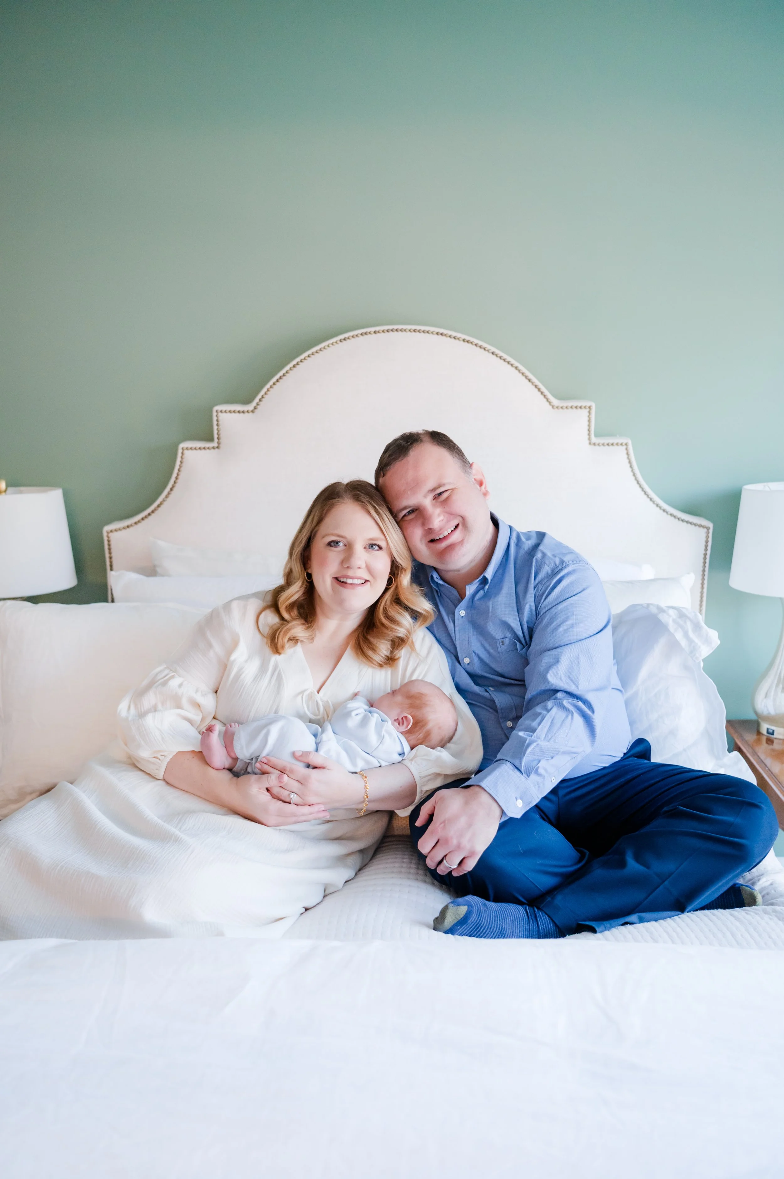 A young couple sitting on a bed, holding a newborn baby, smiling at the camera in a cozy bedroom with green walls and a white headboard. Photo taken in Smyrna. 
