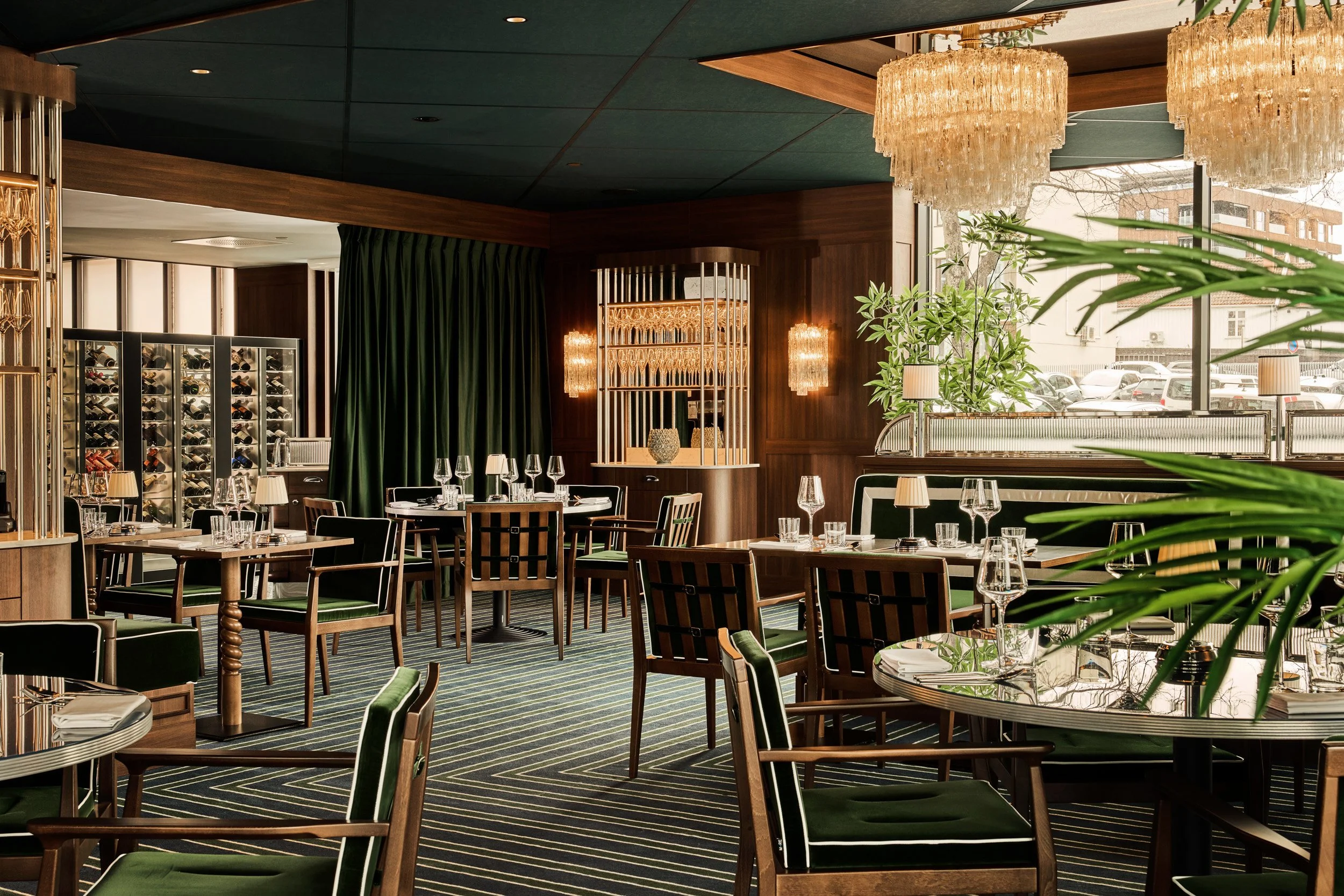 Elegant restaurant interior with wooden furniture, green velvet chairs, and large chandeliers, set for dining with wine glasses and silverware, large windows with a view of parked cars outside.